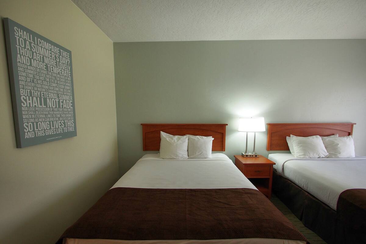 A hotel room with two beds, a nightstand with a lamp, and a wall-mounted light fixture.