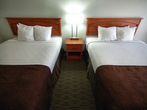 A hotel room with two beds, each with white linens and brown headboards. There is a nightstand with a lamp between the beds. - Stratford Court Hotel (Cedar City)