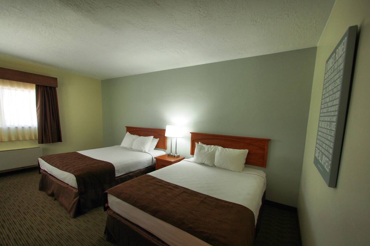 A hotel room with two beds, a nightstand with a lamp, a window with curtains, and a wall-mounted artwork.
