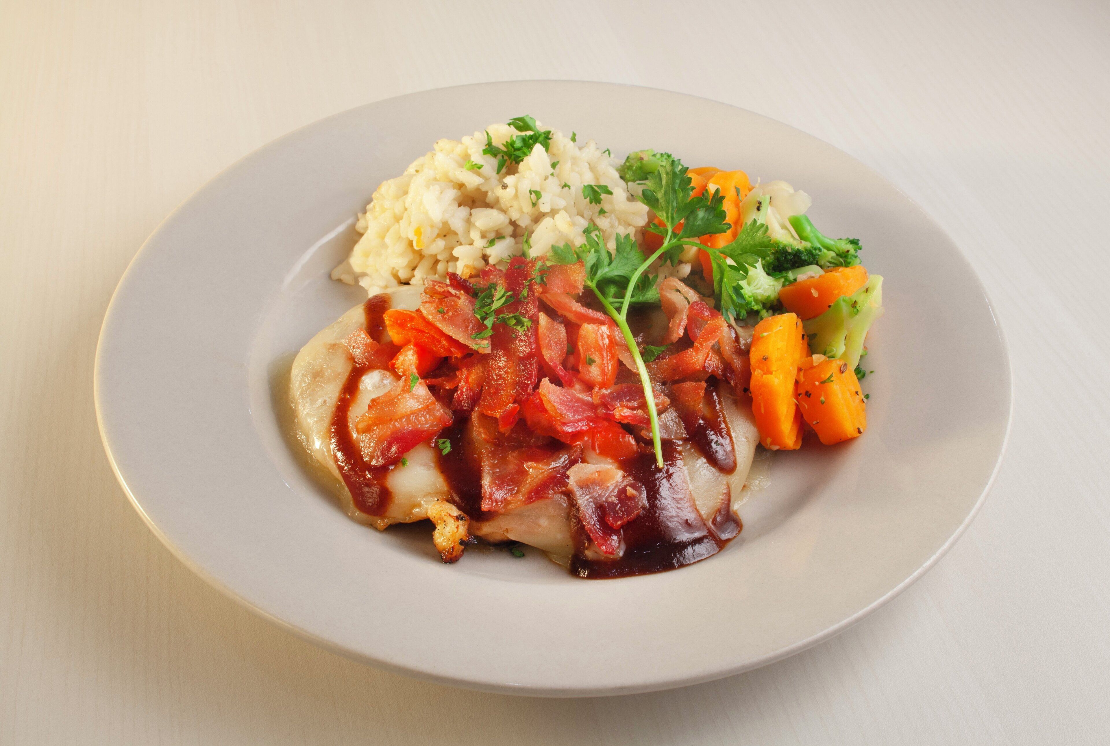 A plate with a piece of meat topped with bacon and tomato sauce, accompanied by rice and mixed vegetables.