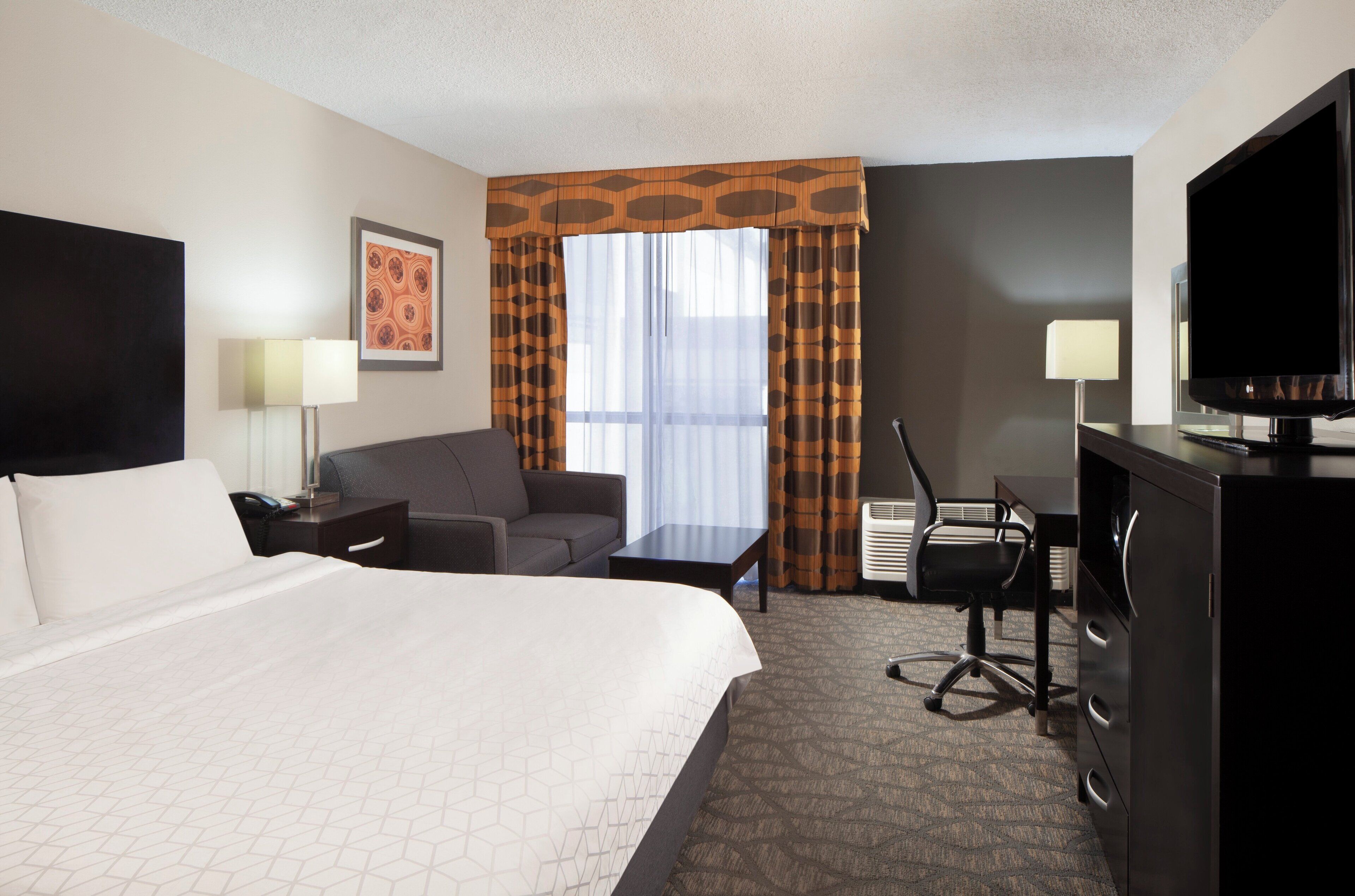 A hotel room with a large bed, a desk with a chair, a television, and a window with curtains.