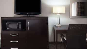 A hotel room with a dark wooden cabinet containing a microwave, a flat-screen TV, and a lamp. There is a chair and a table with a lamp.