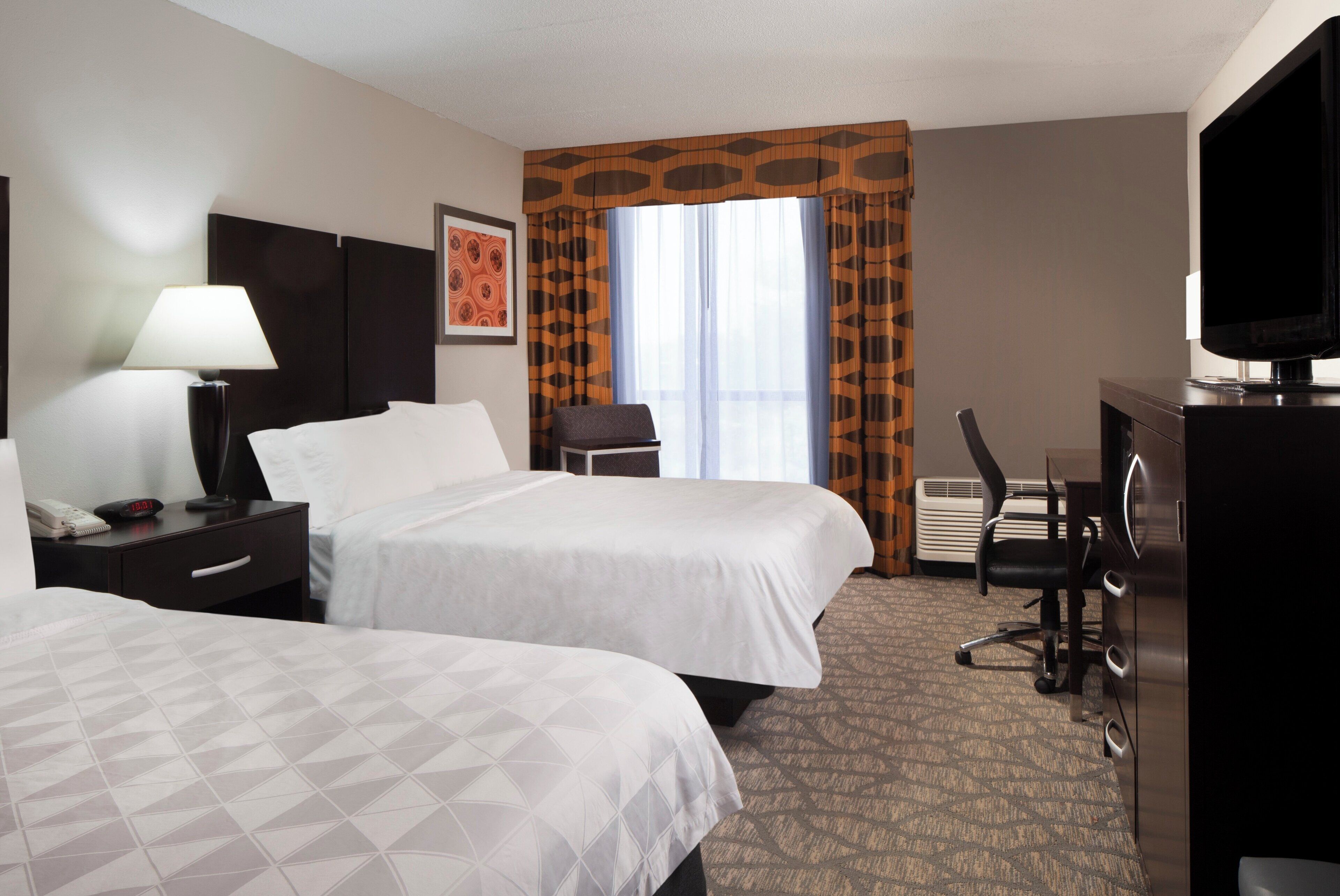 A hotel room with two beds, a desk, a chair, a TV, and a window with curtains.