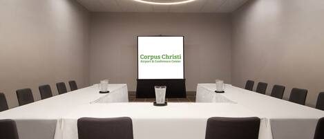 A conference room with a long table, chairs, a projector screen displaying "Corpus Christi Airport & Conference Center", and a patterned carpet.