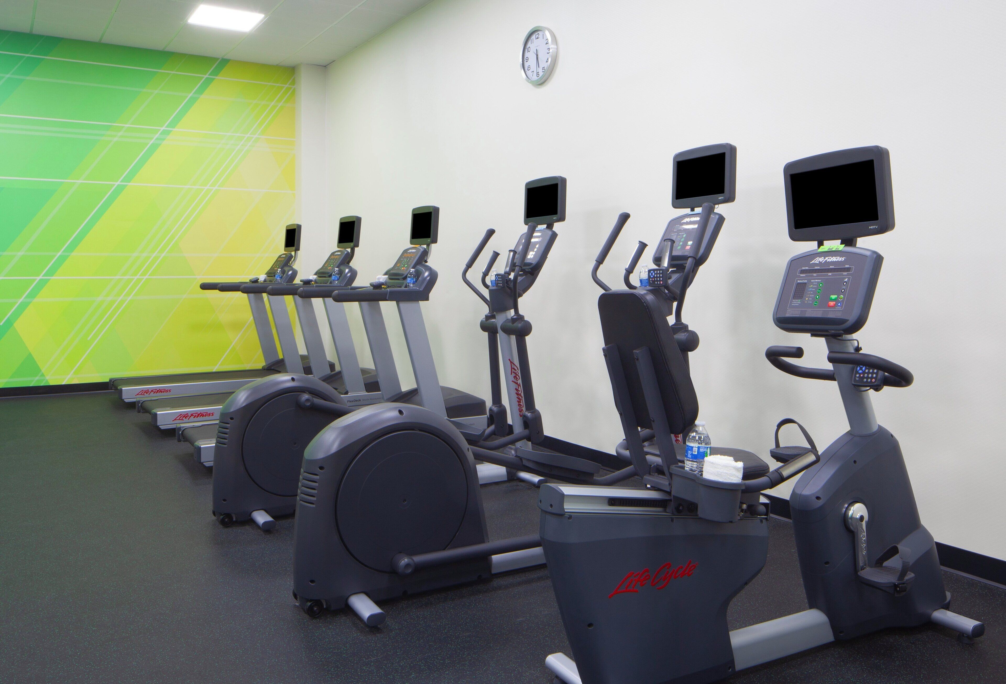 Une rangée de vélos d’appartement dans une salle de sport, avec un mur vert et blanc, une horloge et une bouteille d’eau posée sur l’un des vélos.