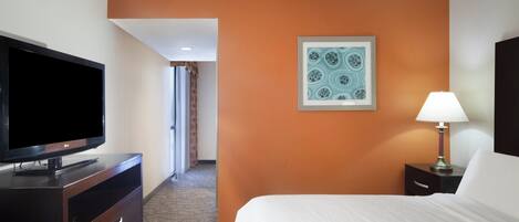 A hotel room with a bed, a TV, a desk, a lamp, and a framed artwork on the wall.