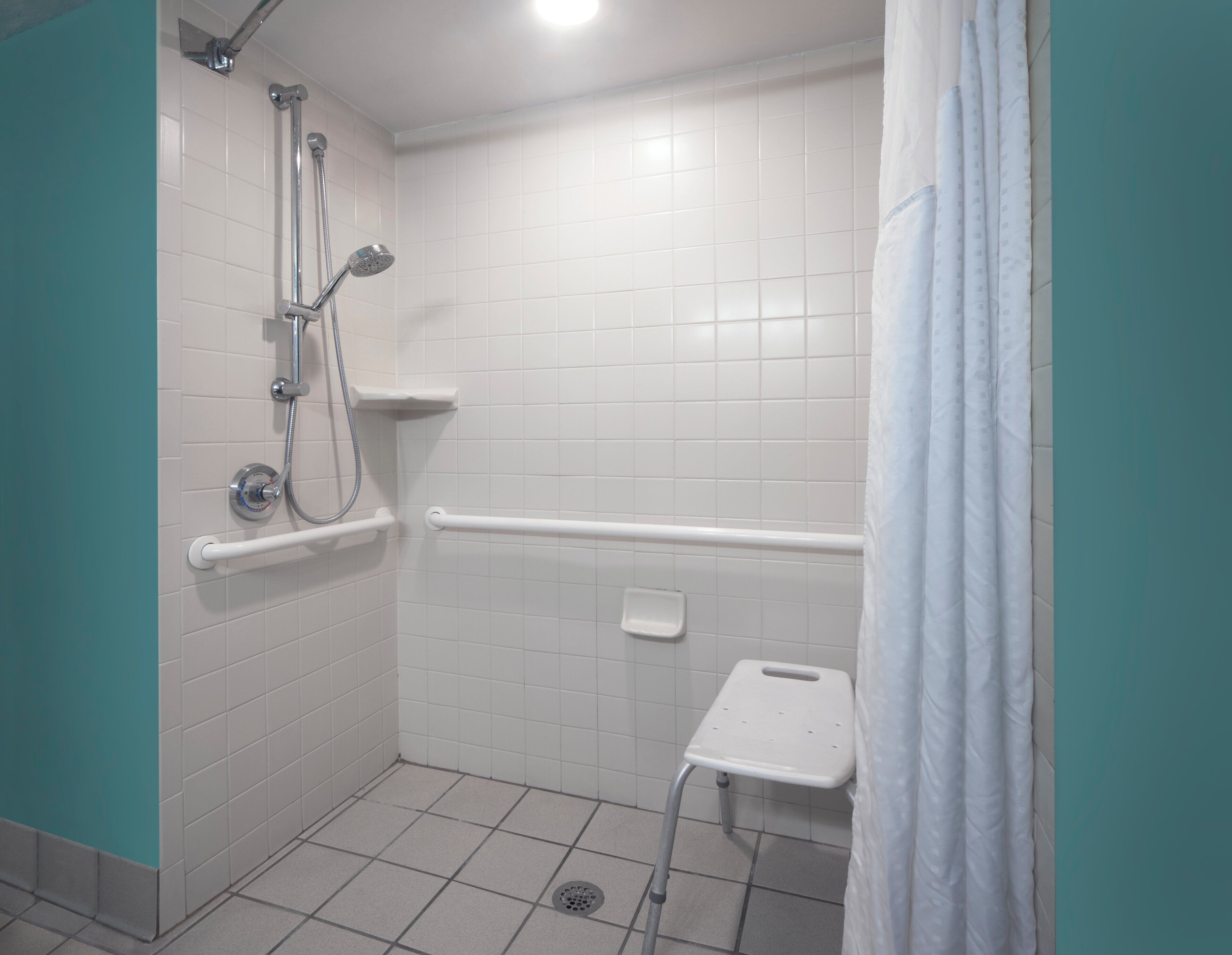 A bathroom with white tiles, a shower with a handheld showerhead, a grab bar, and a small white shelf.