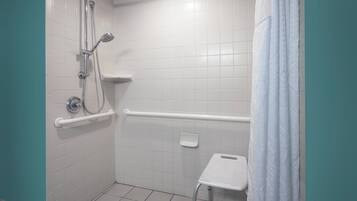 Un baño con azulejos blancos, una ducha con cabezal de ducha de mano, una barra de apoyo y un estante pequeño blanco.