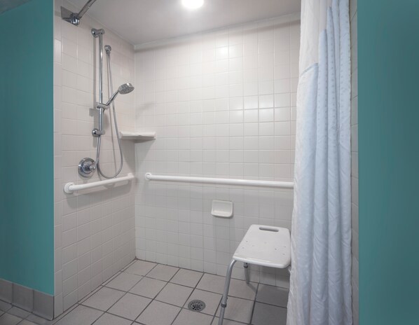 A bathroom with white tiles, a shower with a handheld showerhead, a grab bar, and a small white shelf.