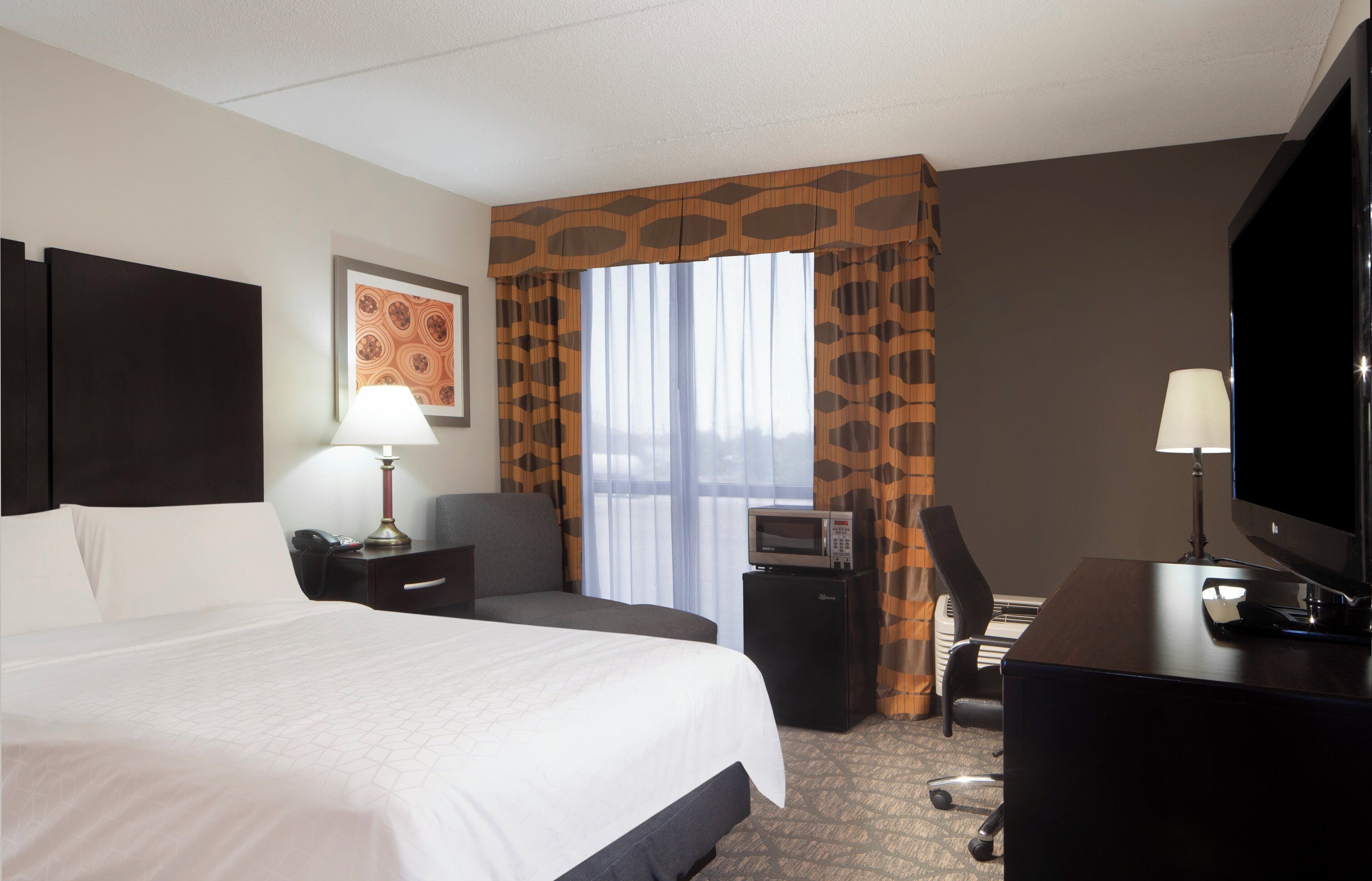 Une chambre d’hôtel avec un grand lit, un bureau avec une télévision, une chaise, une lampe de bureau et une fenêtre avec des rideaux.