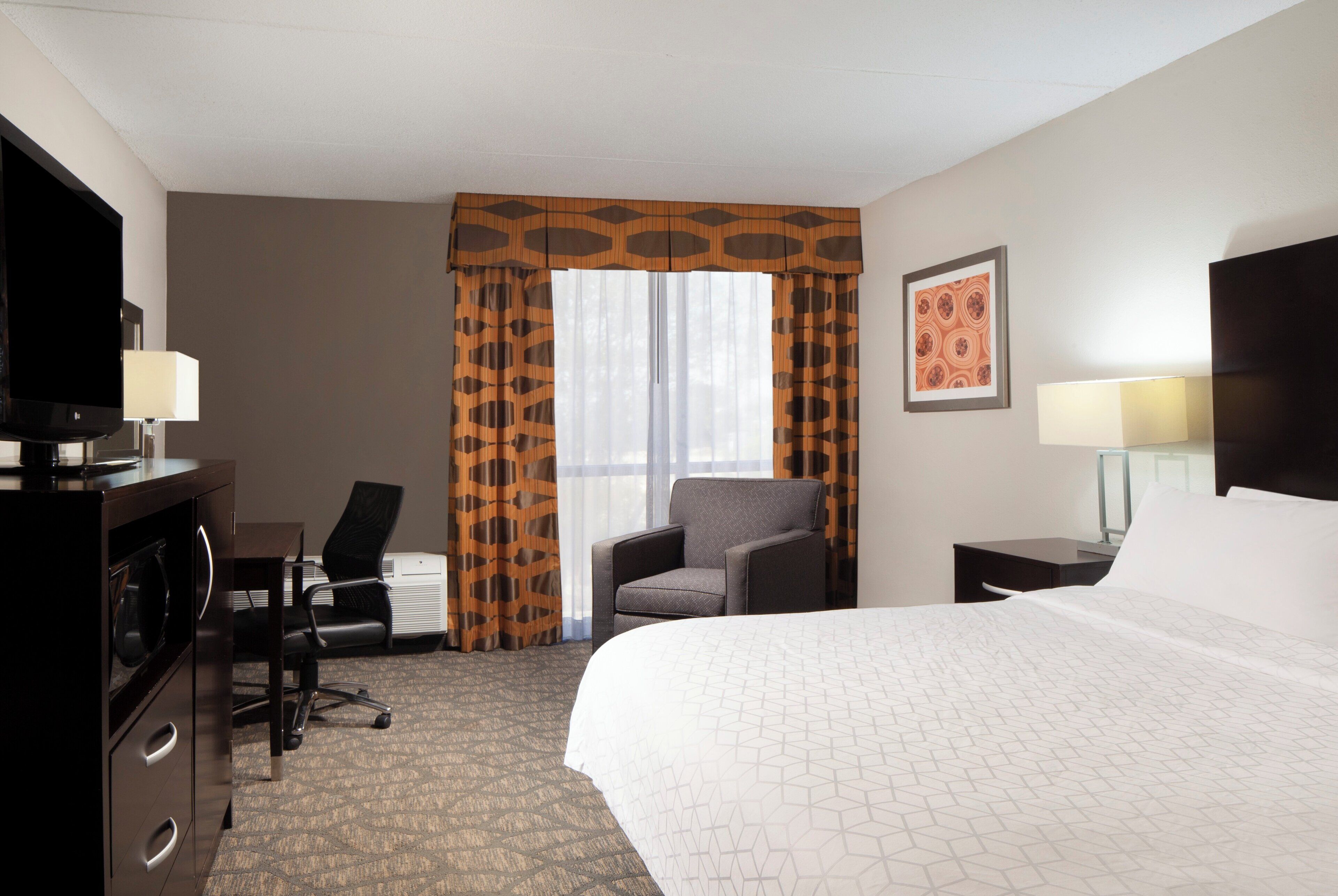 A hotel room with a bed, a desk with a chair, a television, and a window with curtains.