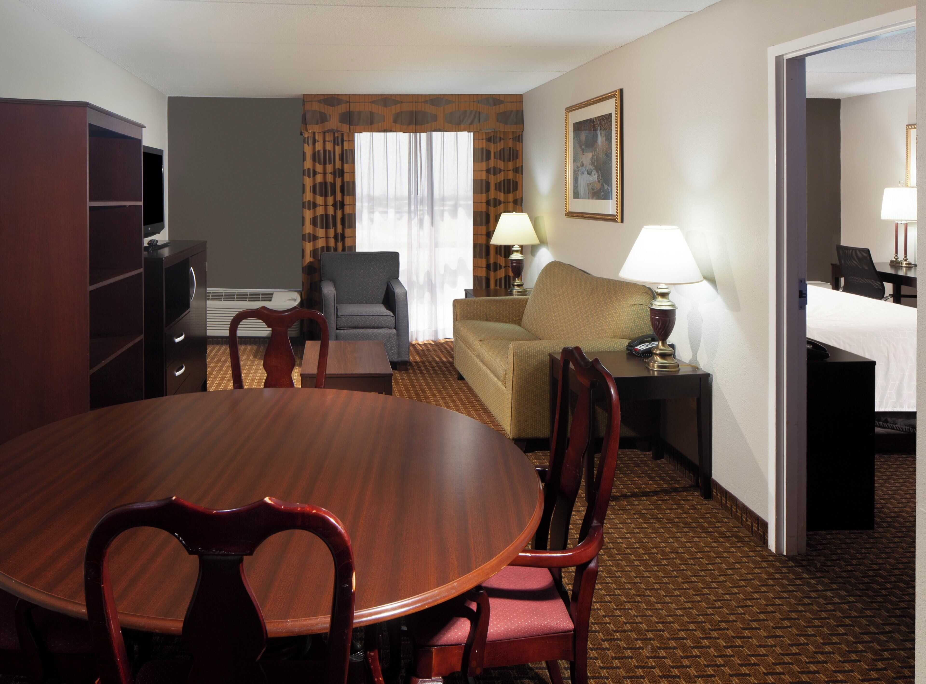 A hotel room with a wooden dining table, two chairs, a sofa, a TV, a lamp, and a bed.