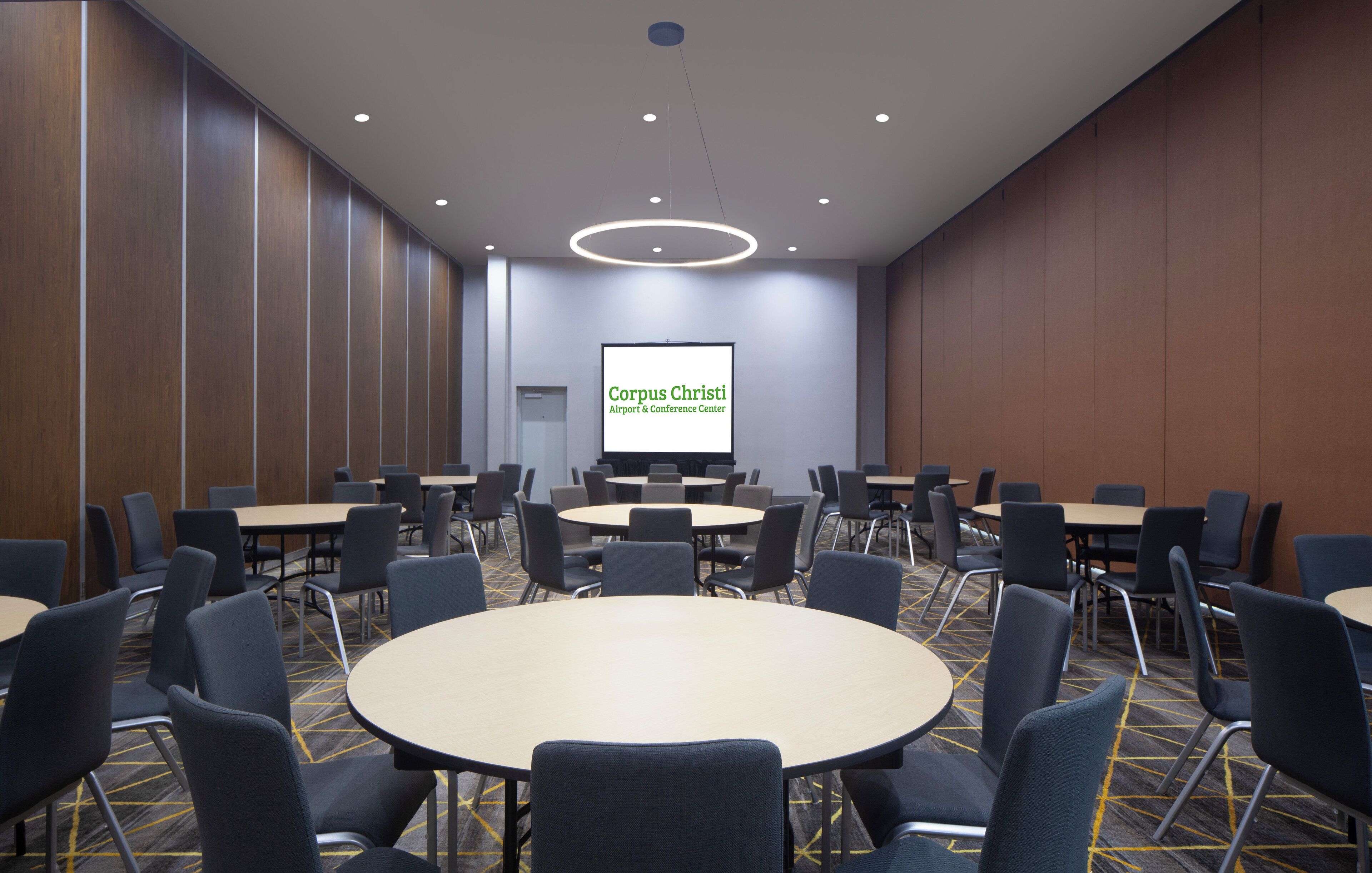 A conference room with round tables and chairs, a large screen displaying 'Corpus Christi Airport & Conference Center', and a modern ceiling design.