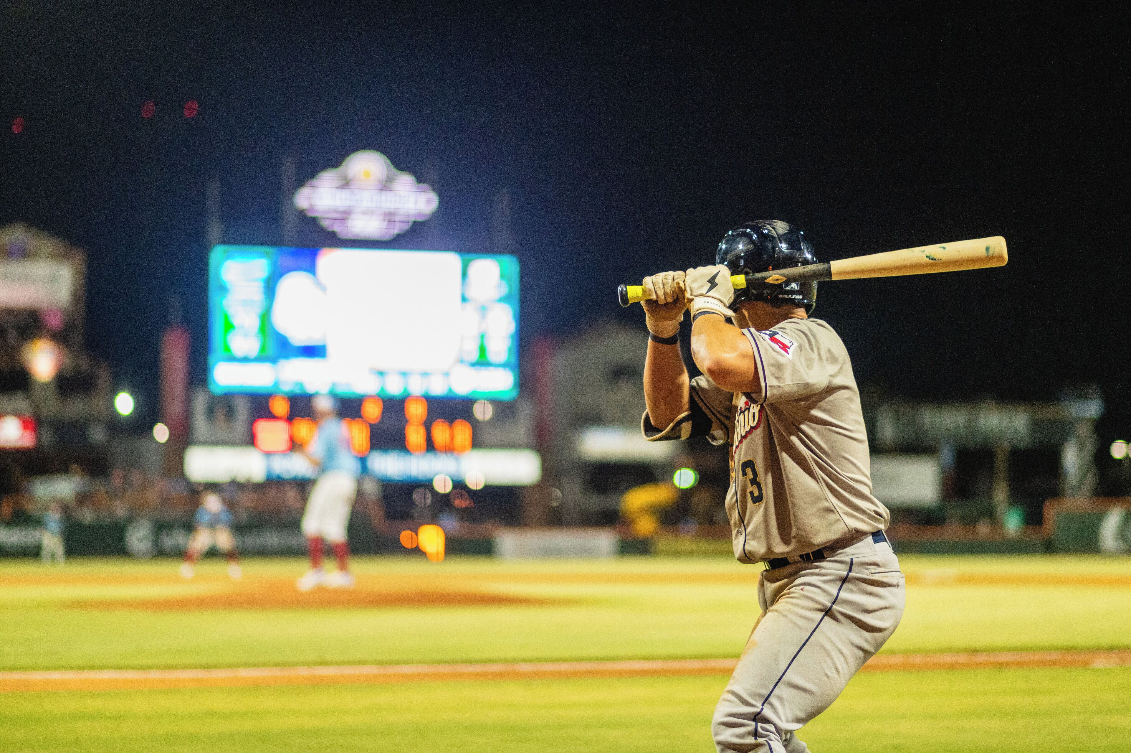 Un jugador de béisbol en un estadio, con un marcador digital grande al fondo.