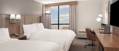 Ein Hotelzimmer mit zwei Betten, einem Schreibtisch, einem Sessel, einem Fernseher und einem Fenster mit Vorhängen.