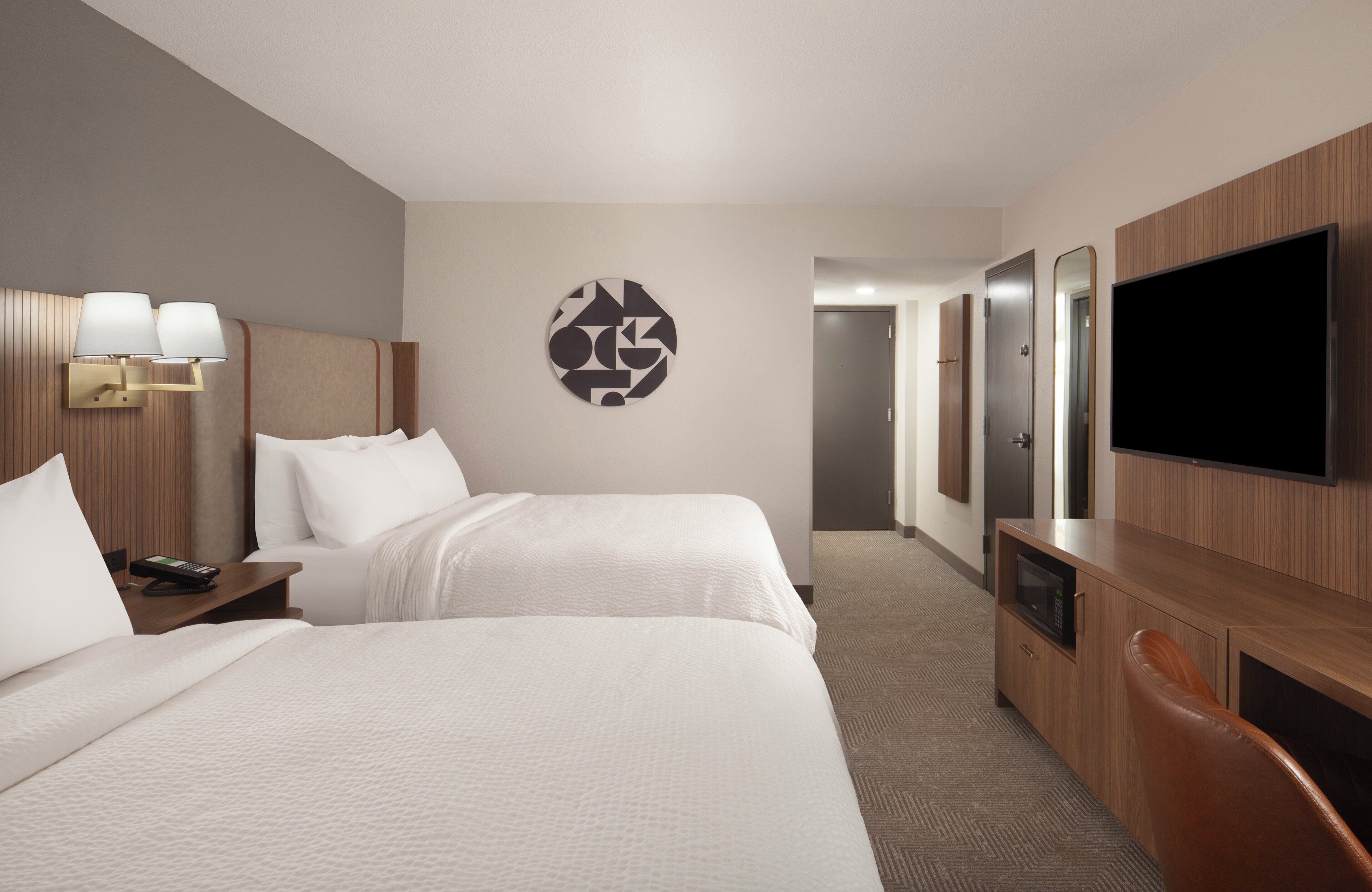A hotel room with two beds, a TV, a desk with a lamp, and a wall art piece.