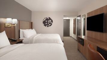 A hotel room with two beds, a TV, a desk with a lamp, and a wall art piece.