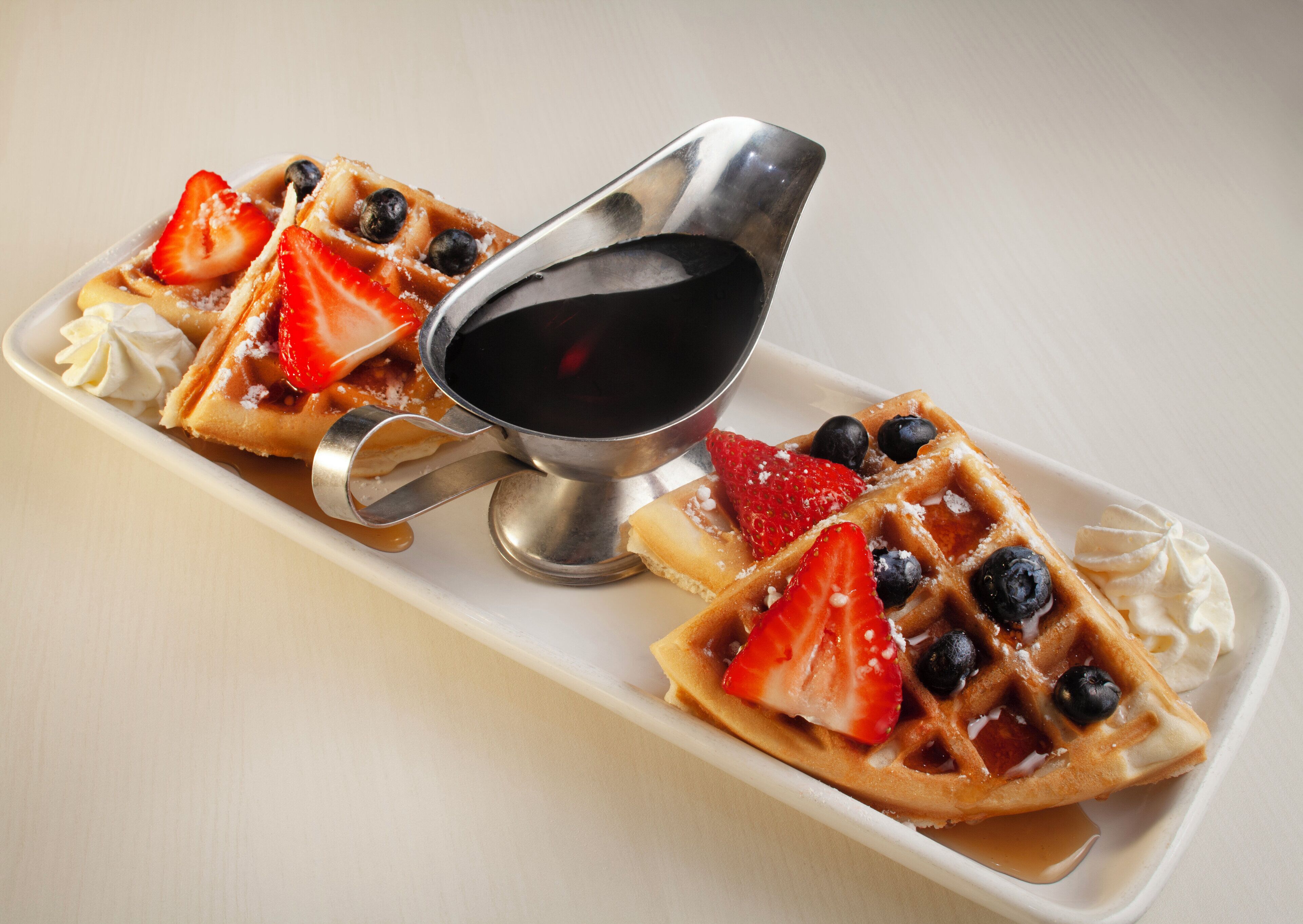 A plate of waffles with strawberries and blueberries, accompanied by a small metal cup of syrup.