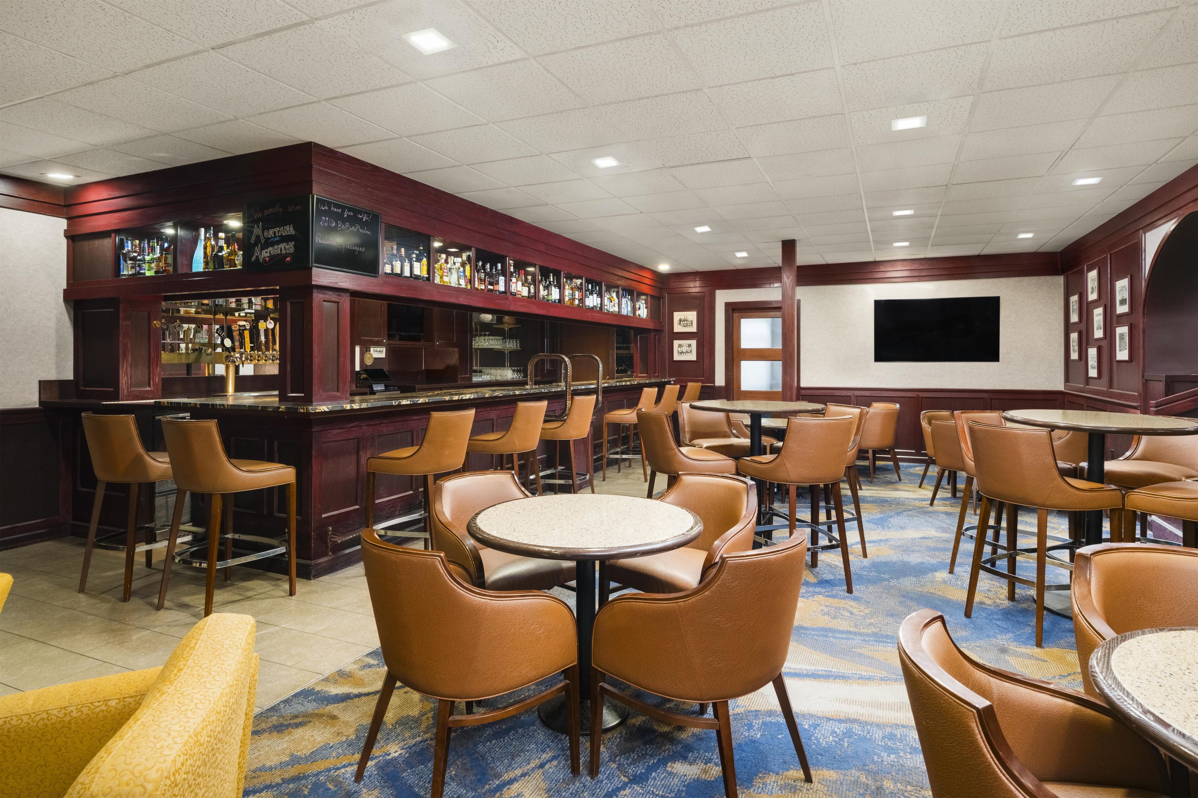 A bar area with multiple seating options including bar stools and tables, a variety of liquor bottles on shelves, and a large screen on the wall.
