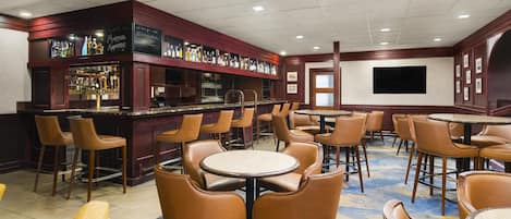 A bar area with multiple seating options including bar stools and tables, a variety of liquor bottles on shelves, and a large screen on the wall.