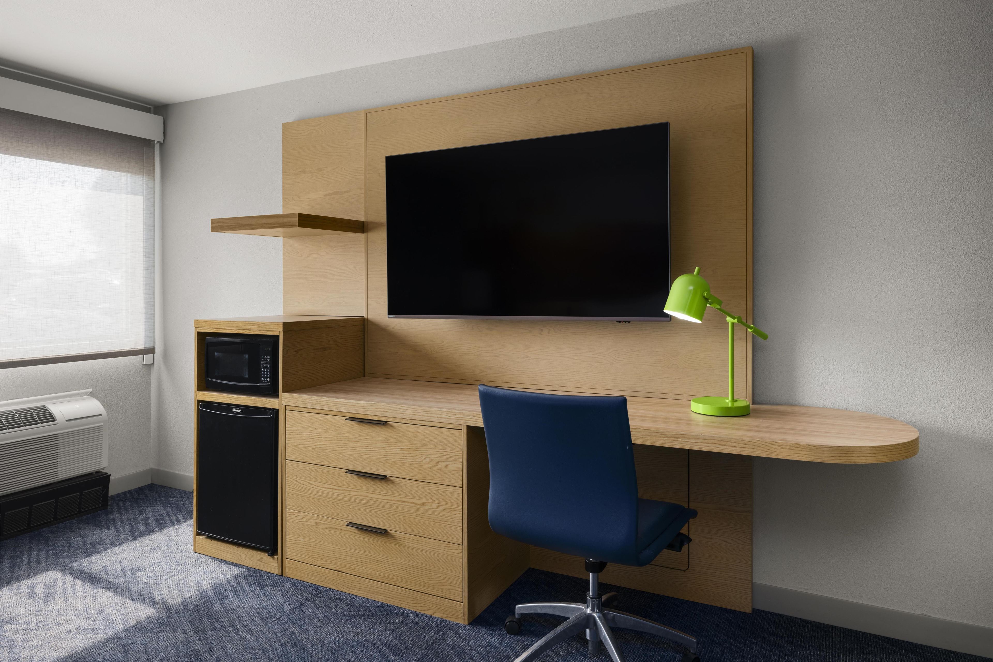 Ein kleines Hotelzimmer mit Schreibtisch, Flachbildfernseher, grüner Schreibtischlampe, Mikrowelle und kleinem Kühlschrank.
