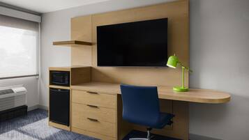 Ein kleines Hotelzimmer mit Schreibtisch, Flachbildfernseher, grüner Schreibtischlampe, Mikrowelle und kleinem Kühlschrank.