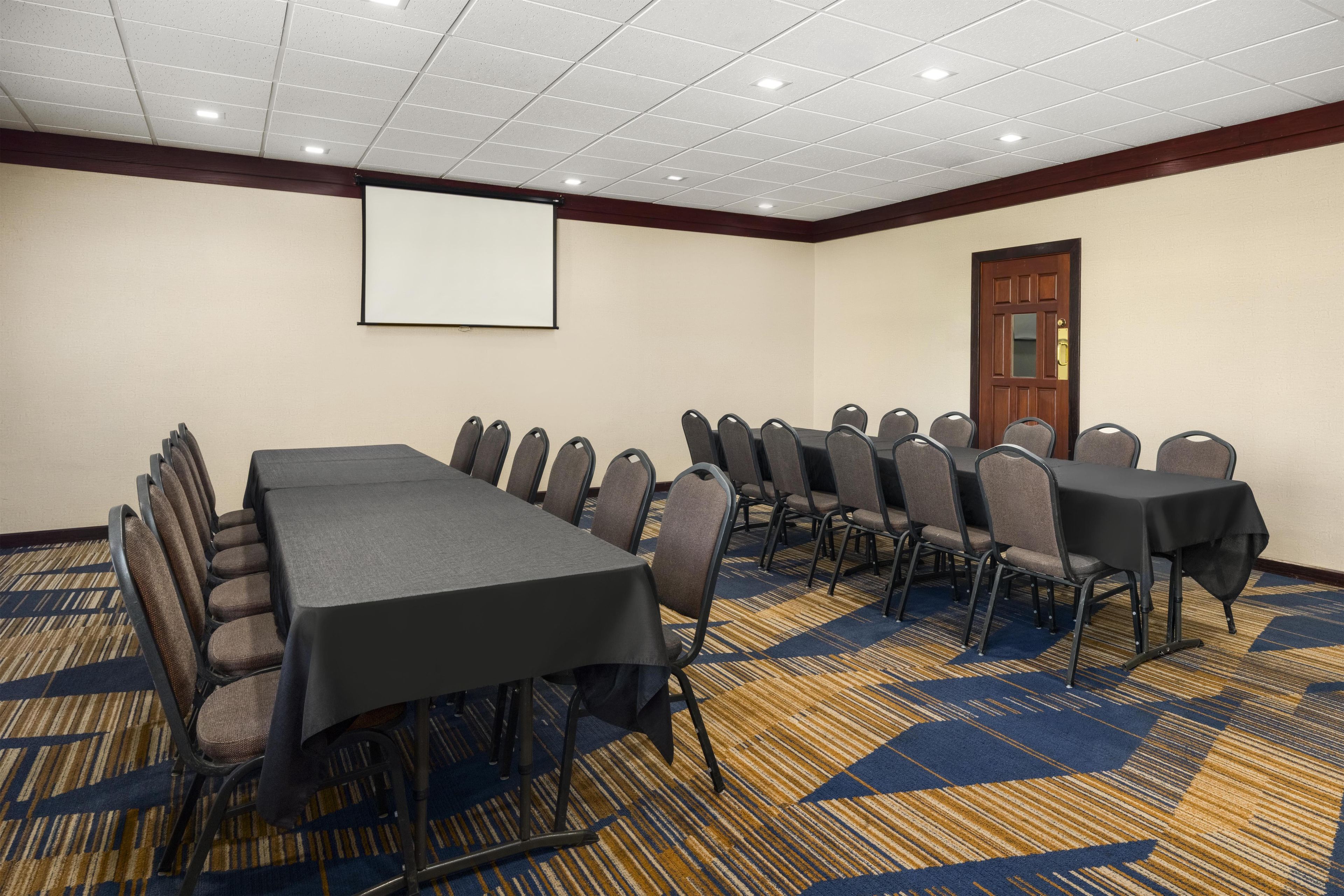 Un salón de conferencias con una mesa larga, varias sillas, una pantalla de proyector y piso alfombrado.