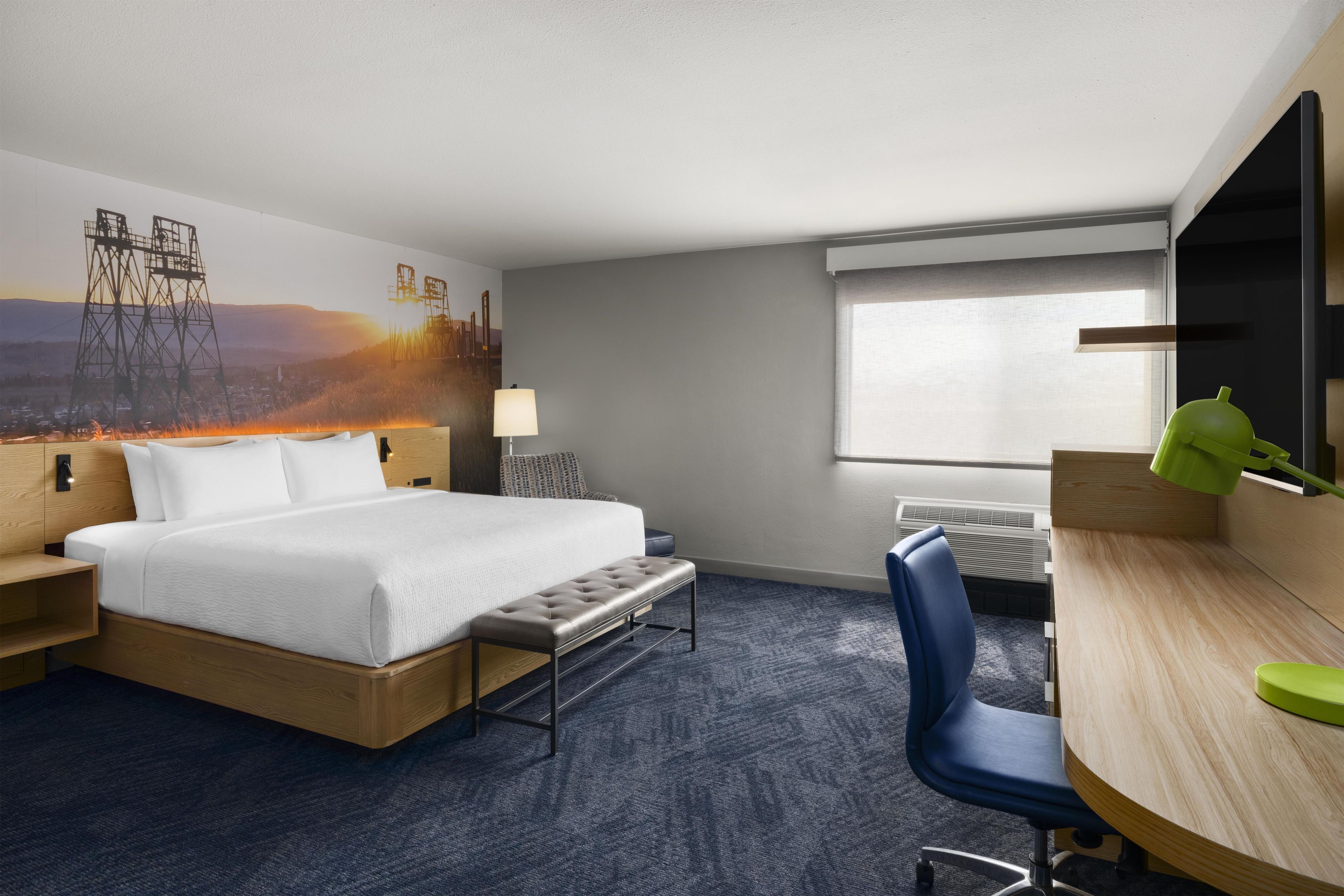 A hotel room with a bed, a desk, a chair, a lamp, and a window.