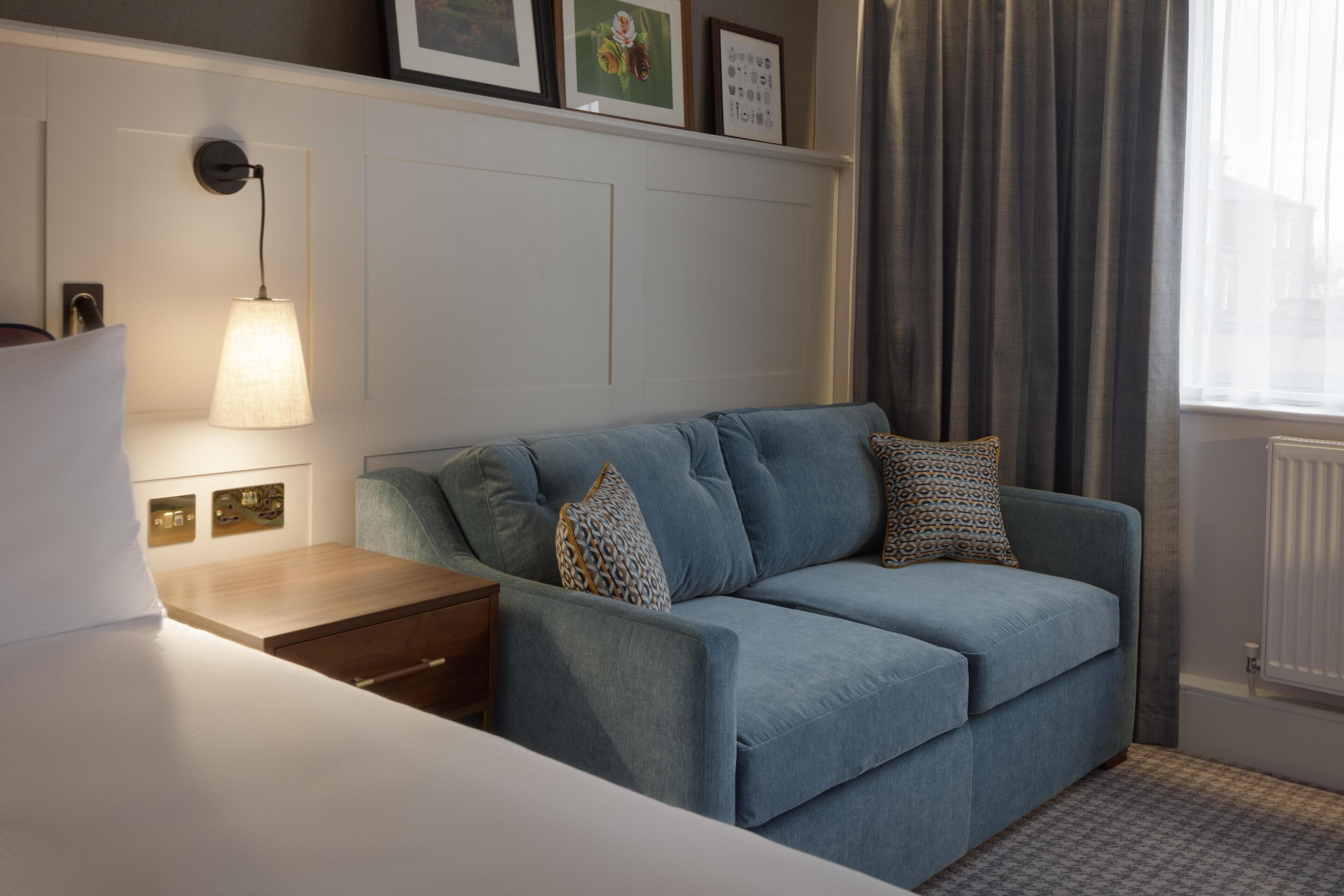 A room with a blue sofa, a bedside table, a lamp, and a window with curtains.