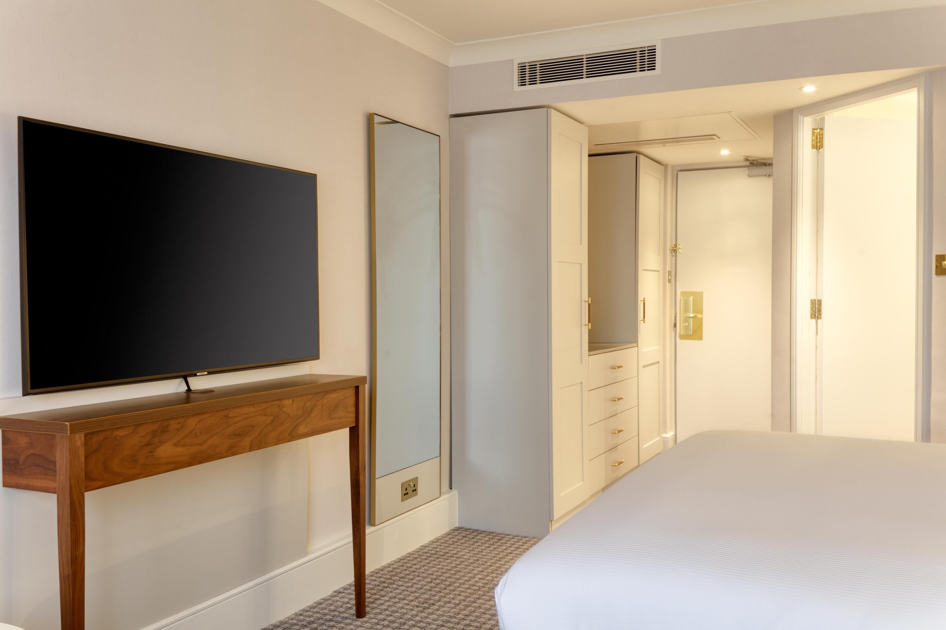Habitación de hotel con cama, televisor sobre una mesa consola de madera, espejo grande, armario y puerta que da a un baño.