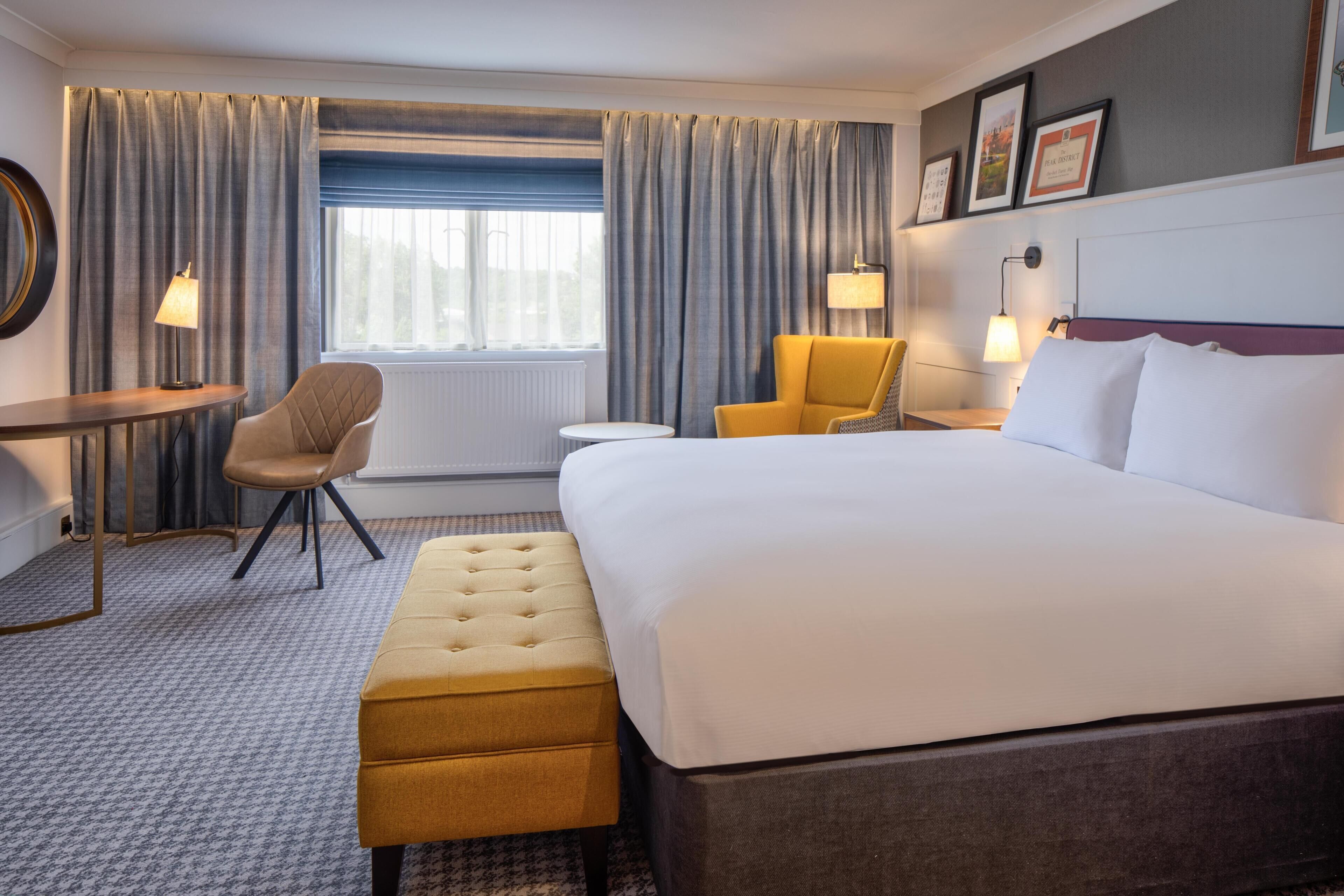 Une chambre d’hôtel avec un grand lit, une petite table ronde, une chaise, un banc et une fenêtre avec des rideaux.