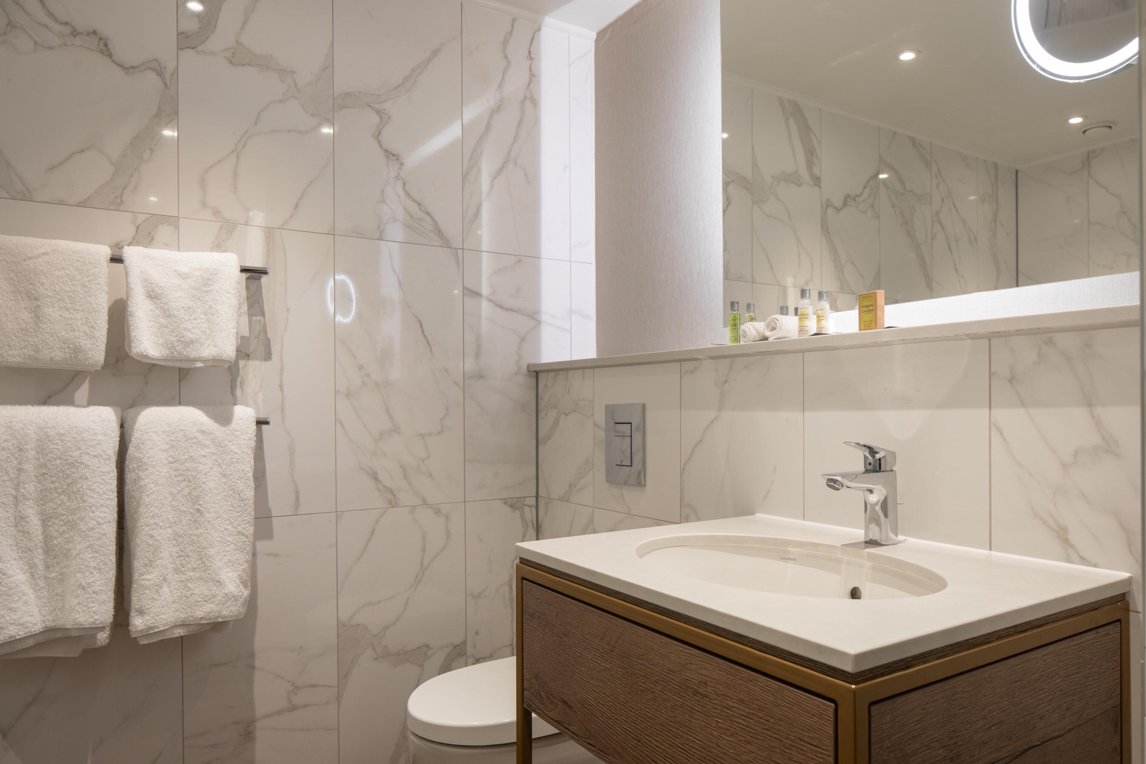 A modern bathroom with a marble wall, a white sink with a wooden vanity, and two white towels on a towel rack.