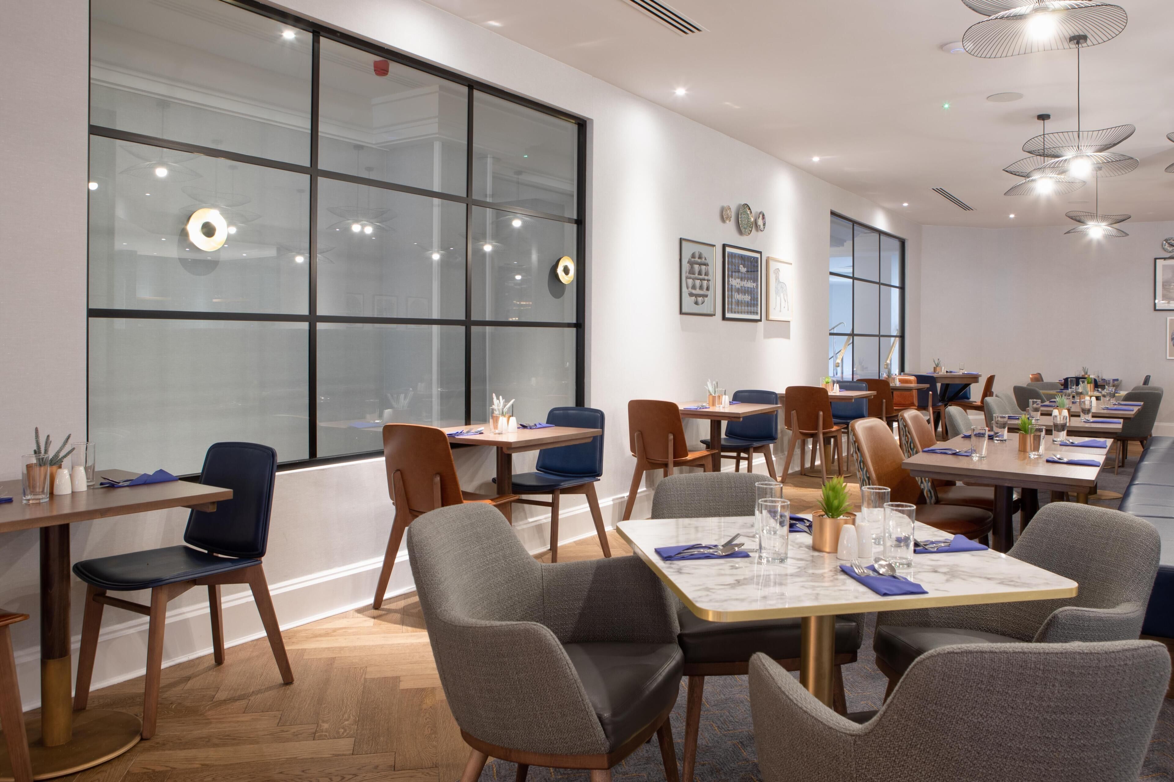 A modern dining area with tables and chairs, large windows, and ceiling lights.
