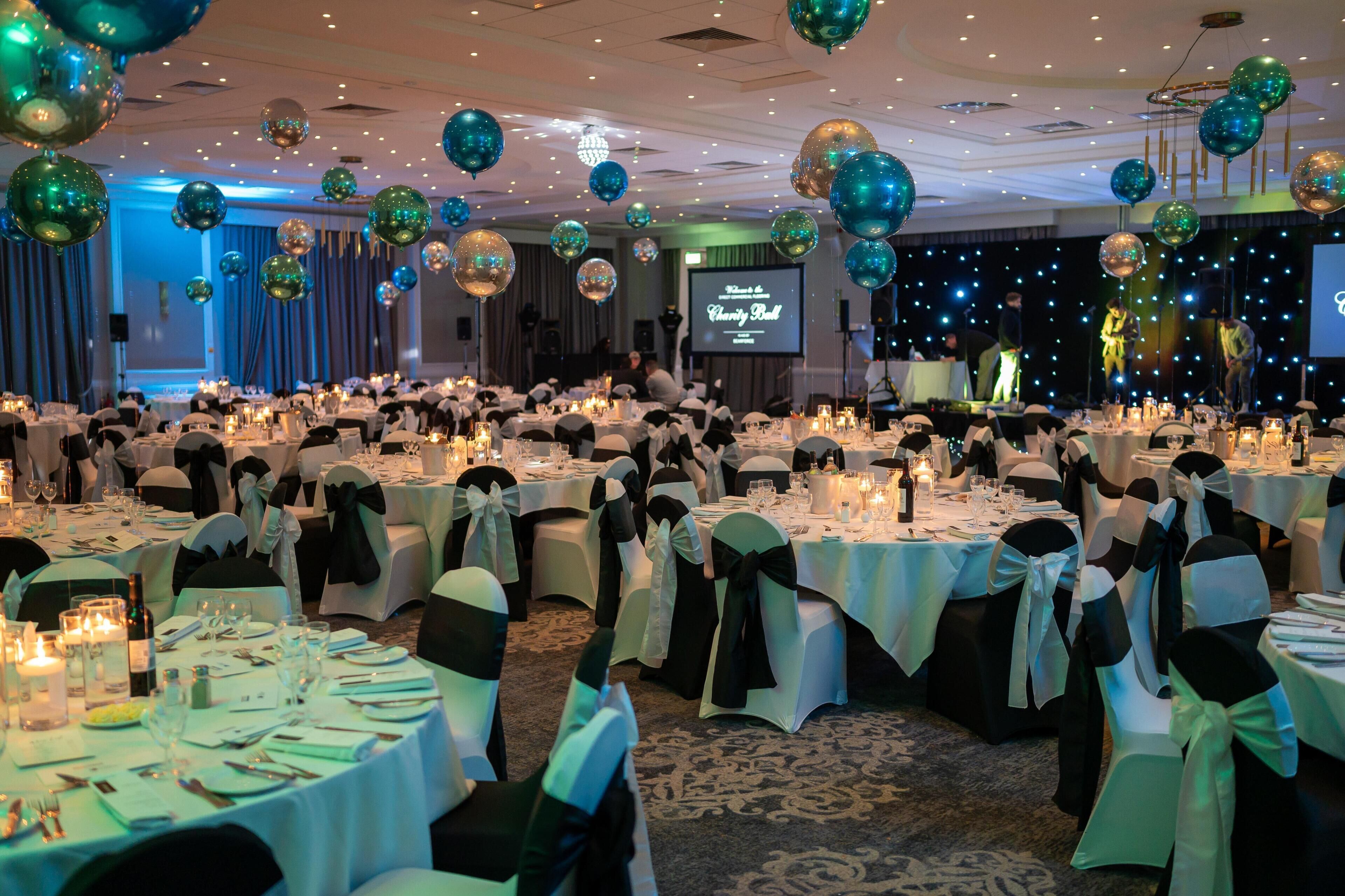Une salle de banquet avec des tables rondes, des chaises noires et blanches, et des ballons aux couleurs turquoise et dorée.