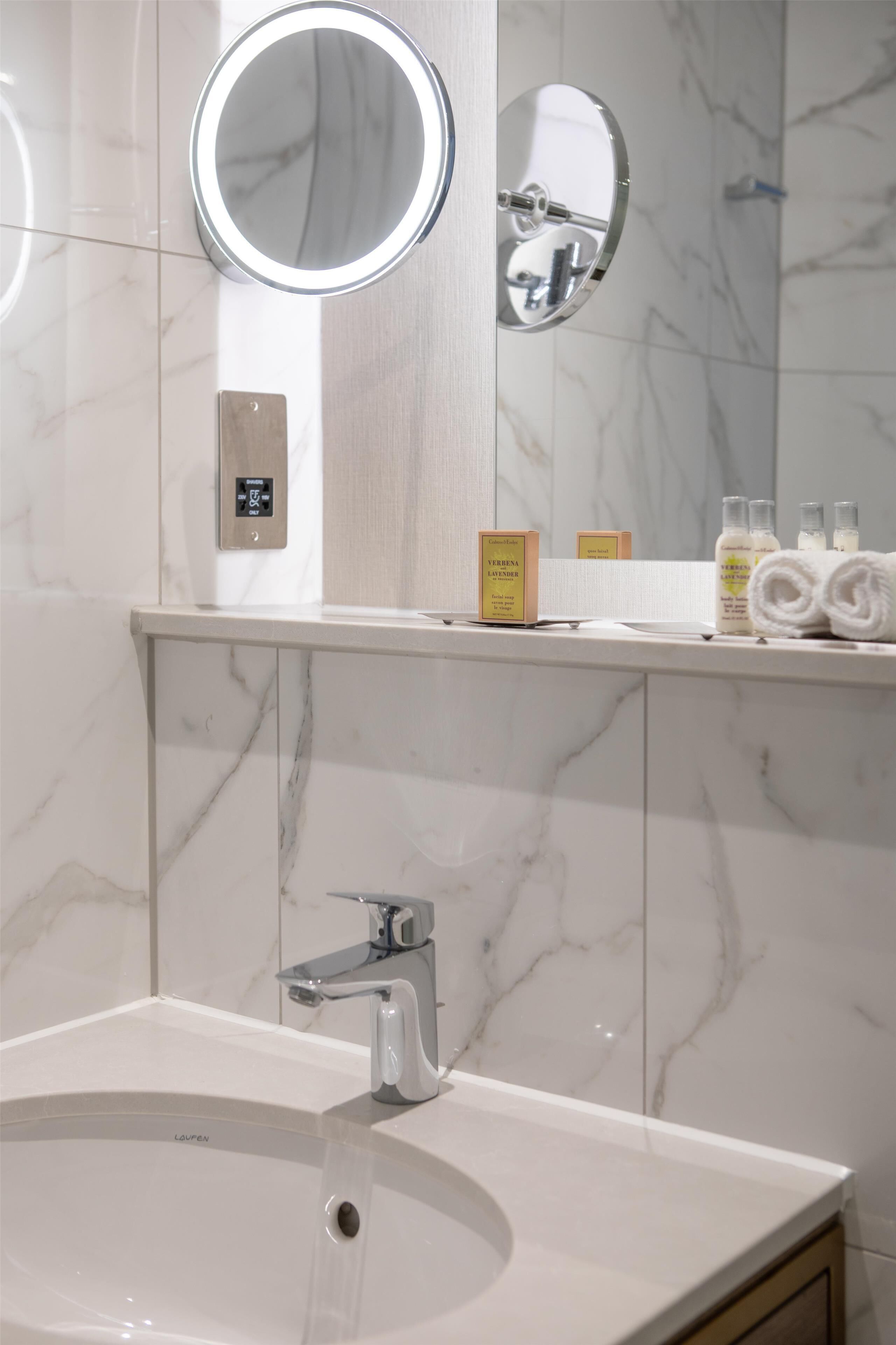 A bathroom with a sink, two mirrors, a towel dispenser, and toiletries.