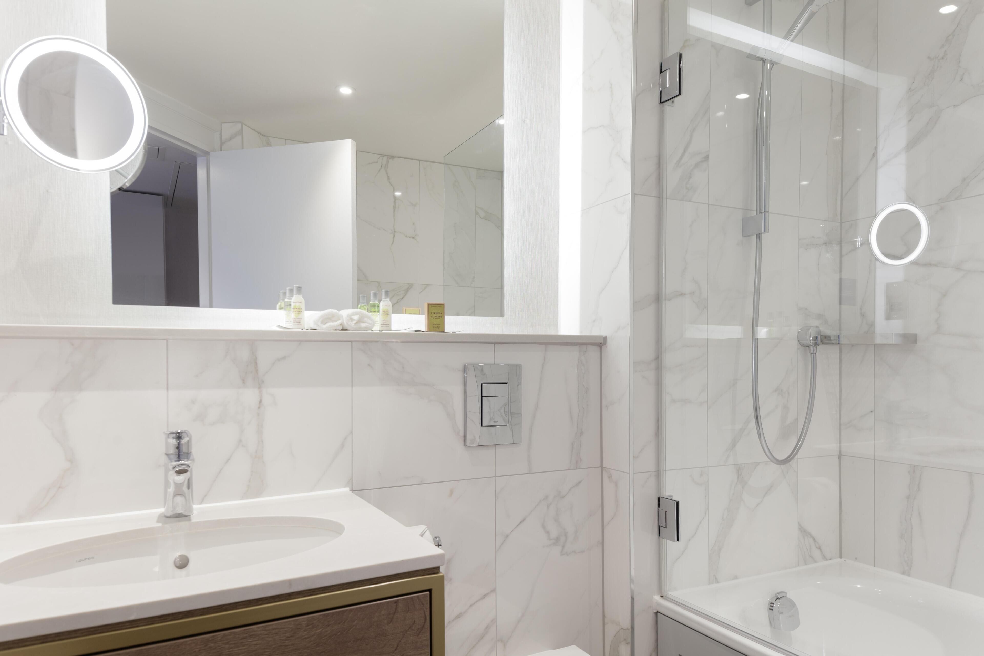 A modern bathroom with a marble sink, a mirror with a light, and a glass-enclosed shower.