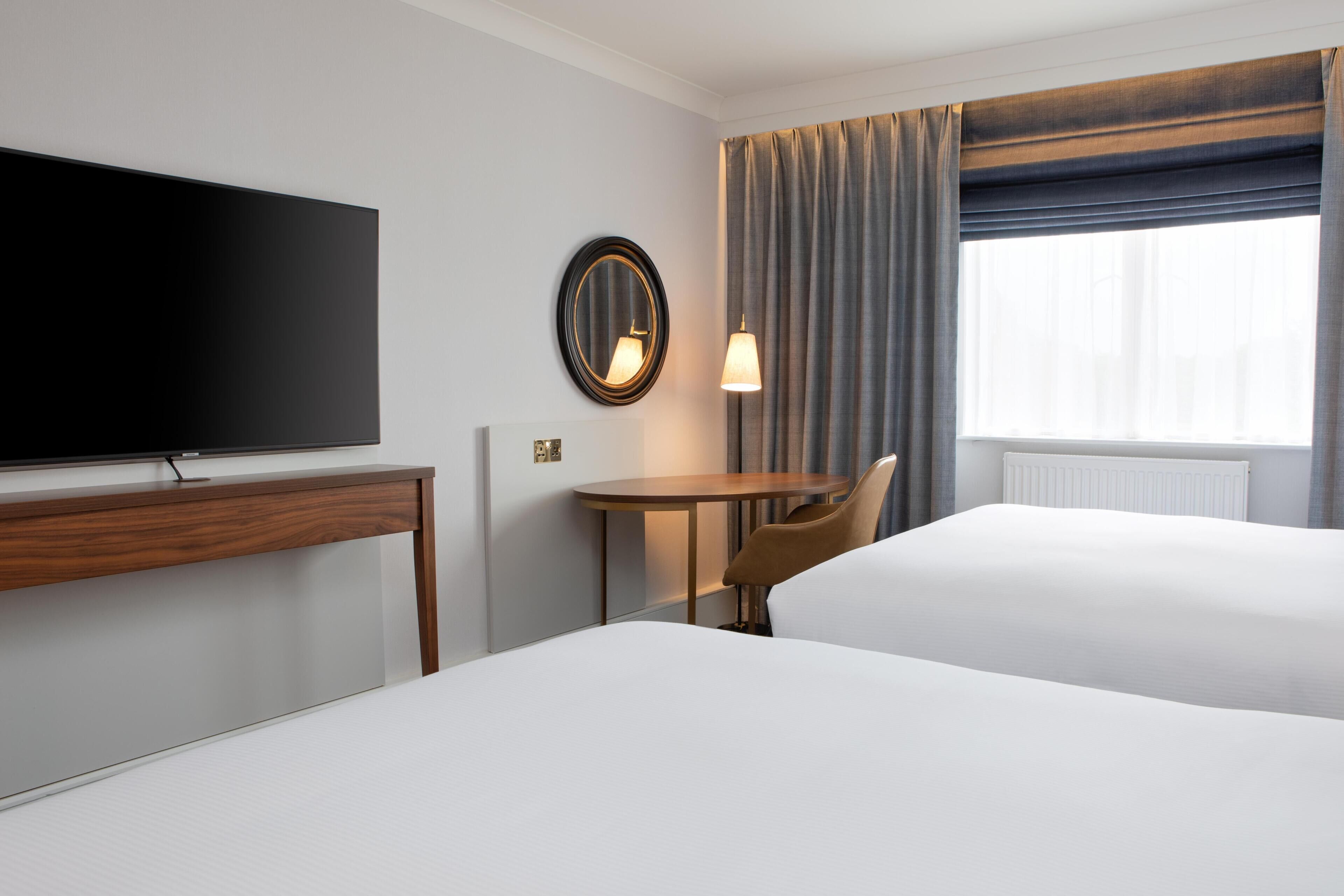 A hotel room with a flat-screen TV, a round mirror, a small table with a lamp, two beds, and a window with curtains.
