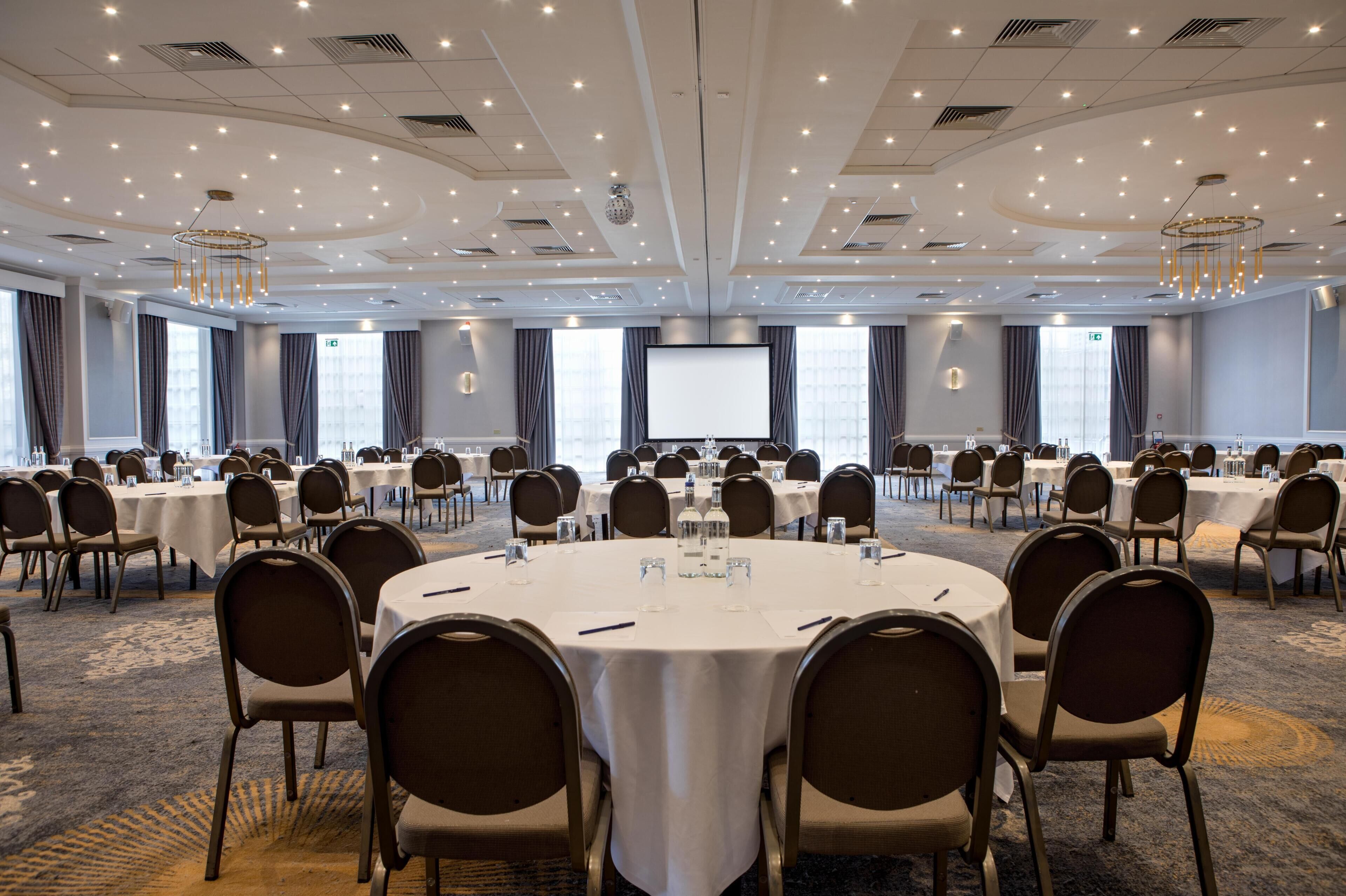A conference room with round tables, chairs, a projector screen, and a carpeted floor.