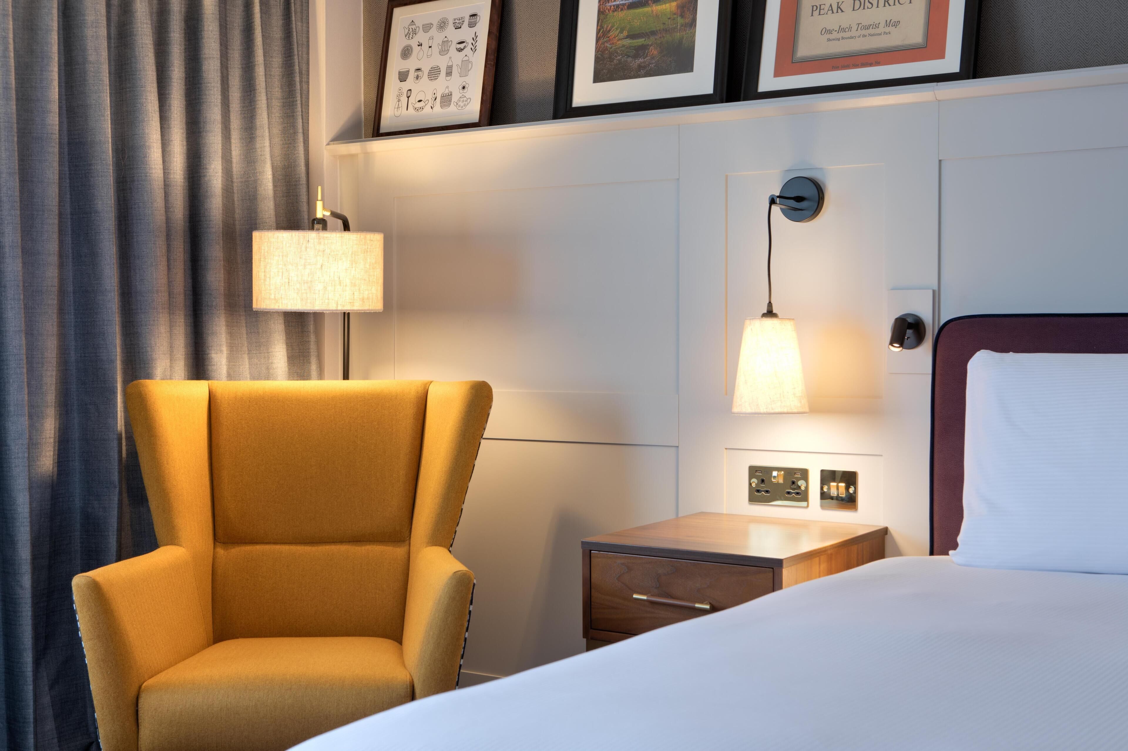 A hotel room with a bed, a chair, a nightstand, a lamp, and framed pictures on the wall.