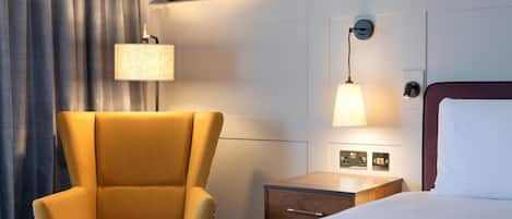 A hotel room with a bed, a chair, a nightstand, a lamp, and framed pictures on the wall.