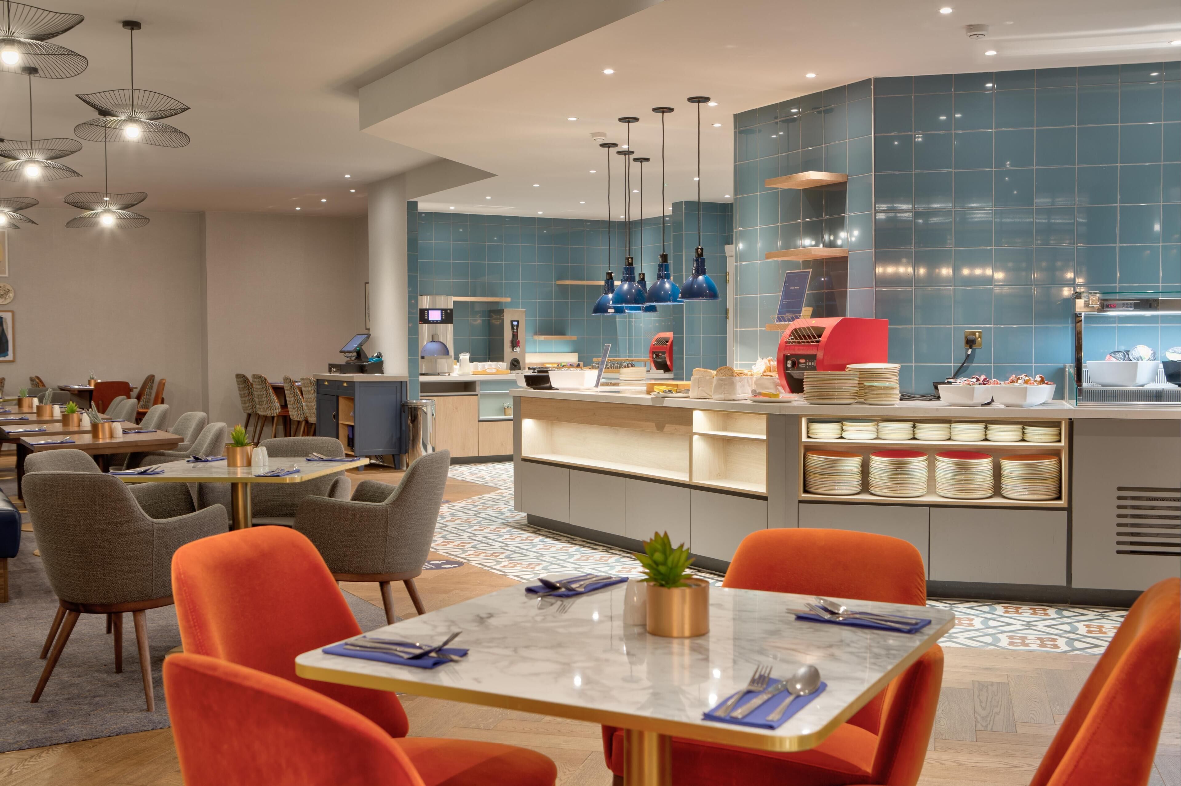 A modern hotel lobby with a central counter, seating area, and a kitchen in the background.