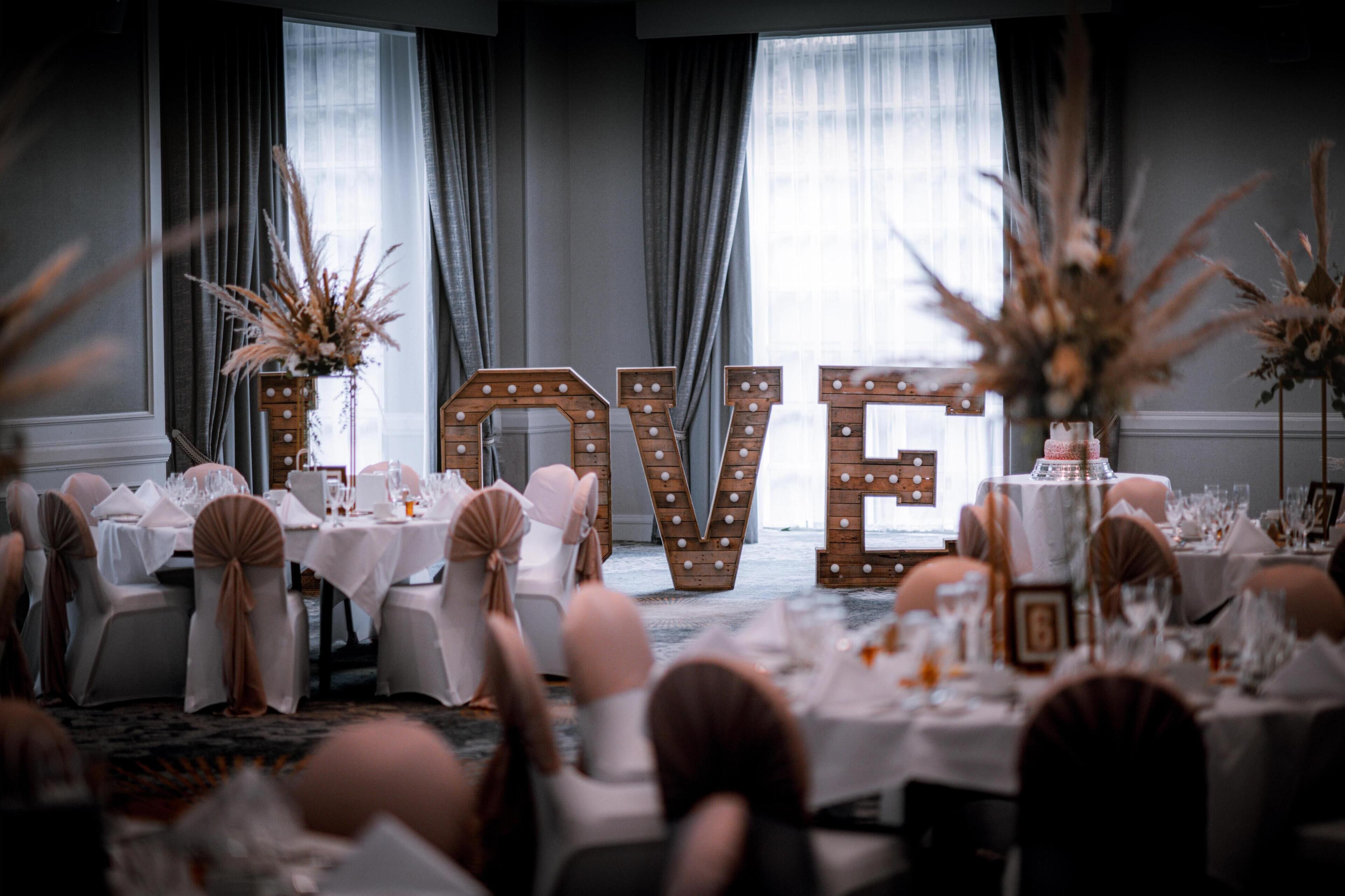 A banquet hall with tables set for a formal event, featuring large illuminated letters spelling 'LOVE' and floral centerpieces.