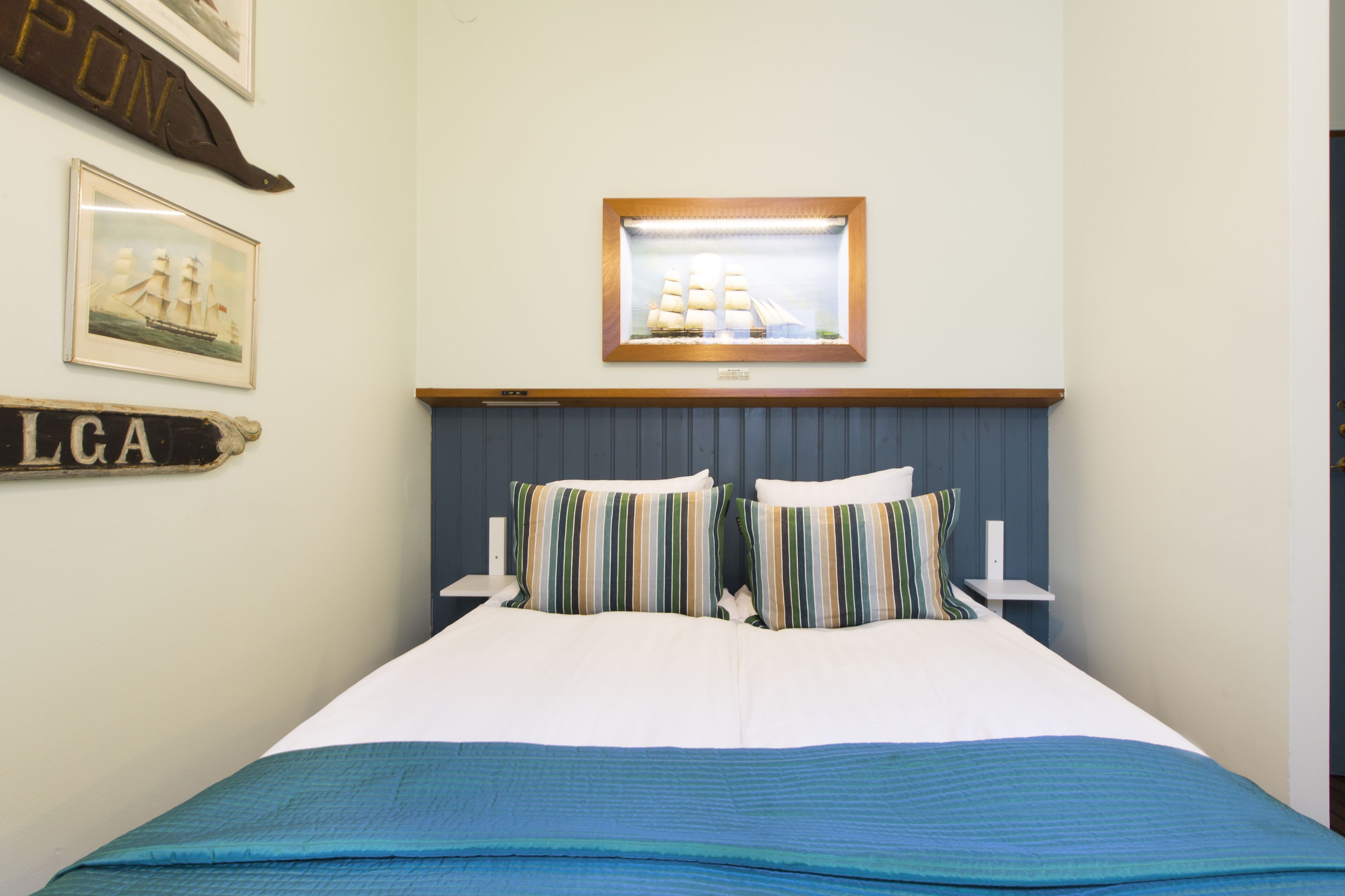 Una cama bien hecha con almohadillas a rayas y una colcha azul, un cuadro con una imagen de un barco en la pared, y un cartel con las letras 