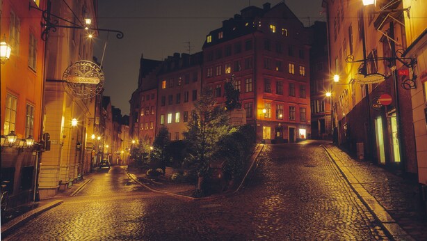 A cobblestone street lined with historic buildings, illuminated by street lamps and featuring a prominent sign for a hotel. - Lord Nelson Hotel (Stockholm)