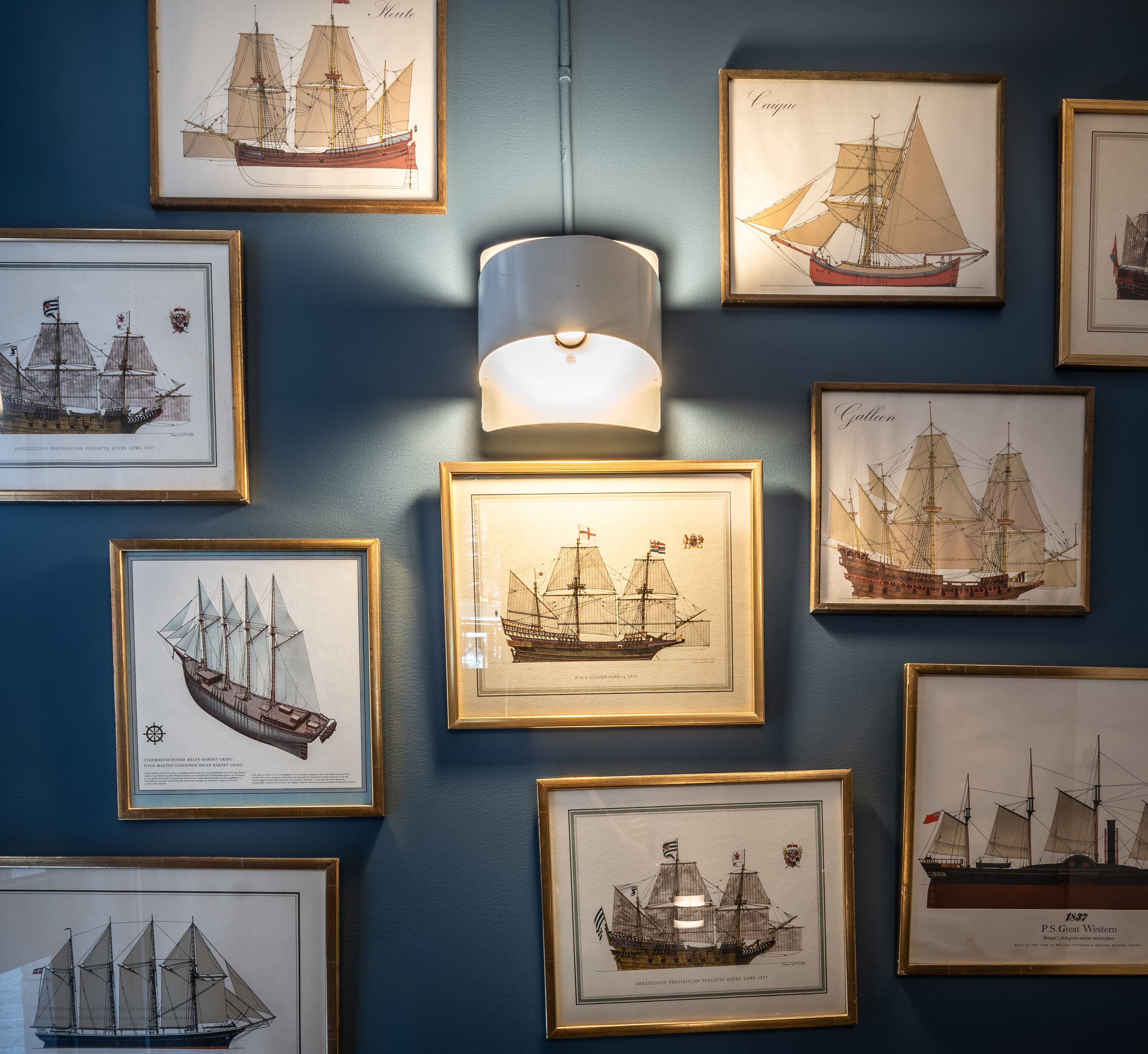 A wall with framed maritime paintings, a hanging light fixture, and a blue wall.