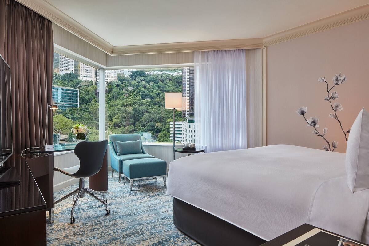 A hotel room with a large bed, a desk, two chairs, and a view of a cityscape and greenery.
