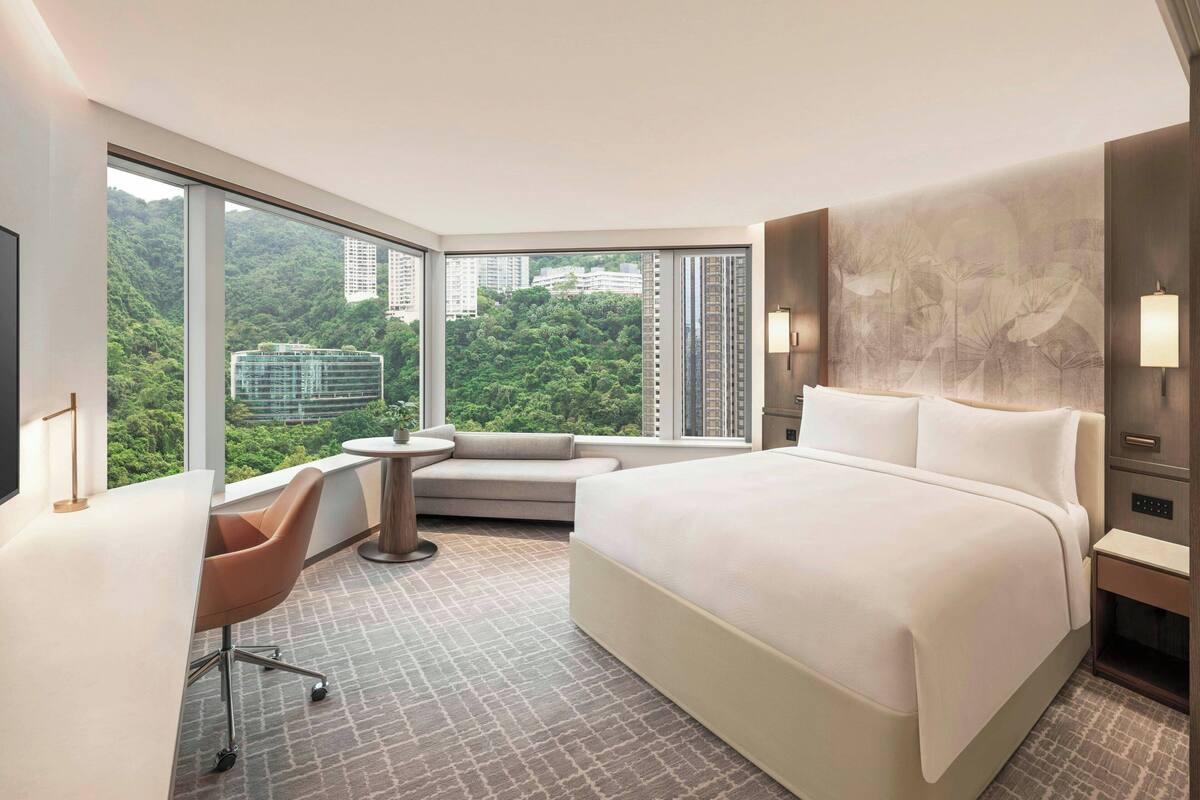A modern hotel room with a large bed, a desk, a chair, and a view of a cityscape through large windows.