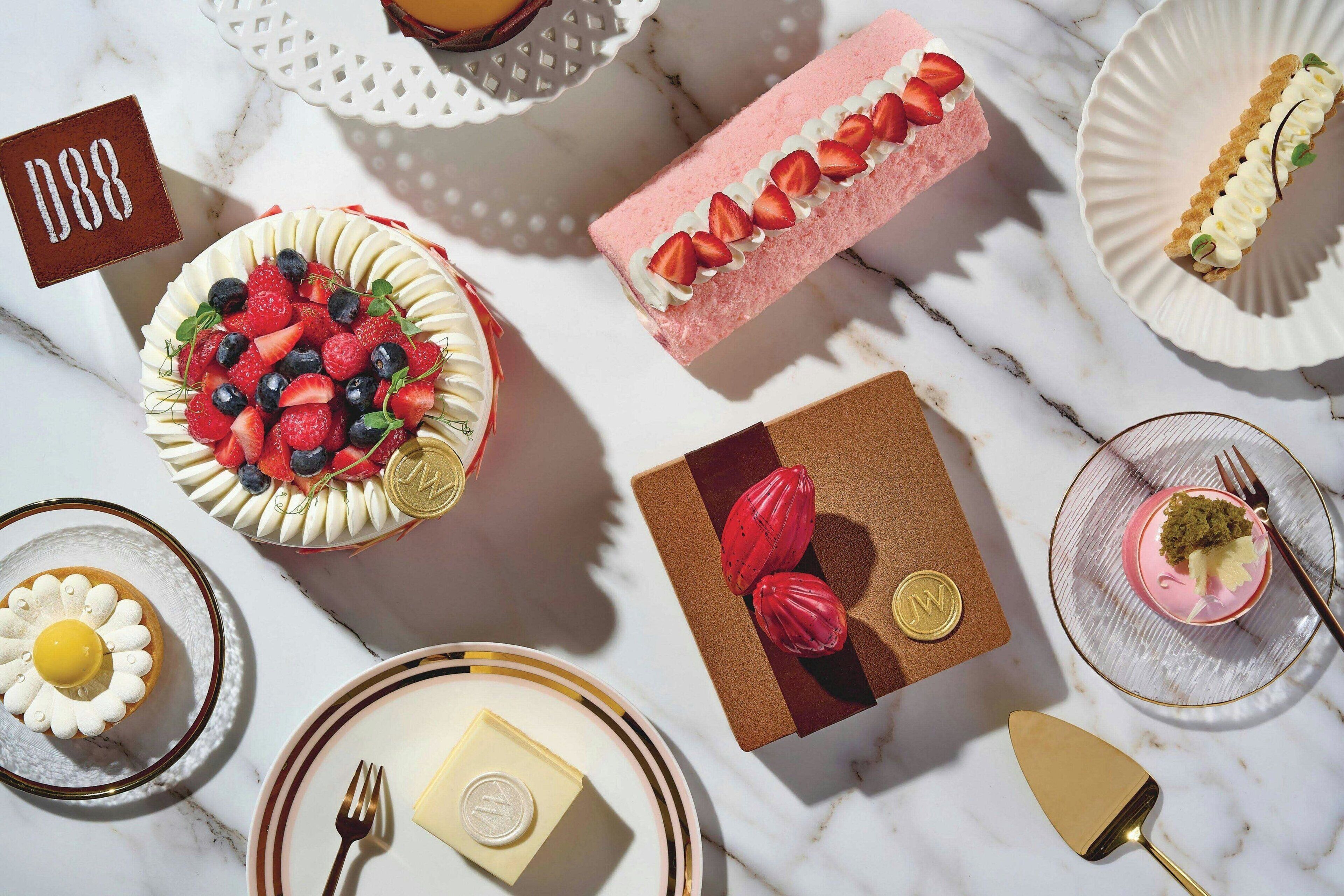 A variety of desserts on a marble table, including a cake with strawberries and blueberries, a pink cake with strawberries, and a tart with whipped cream and strawberries.