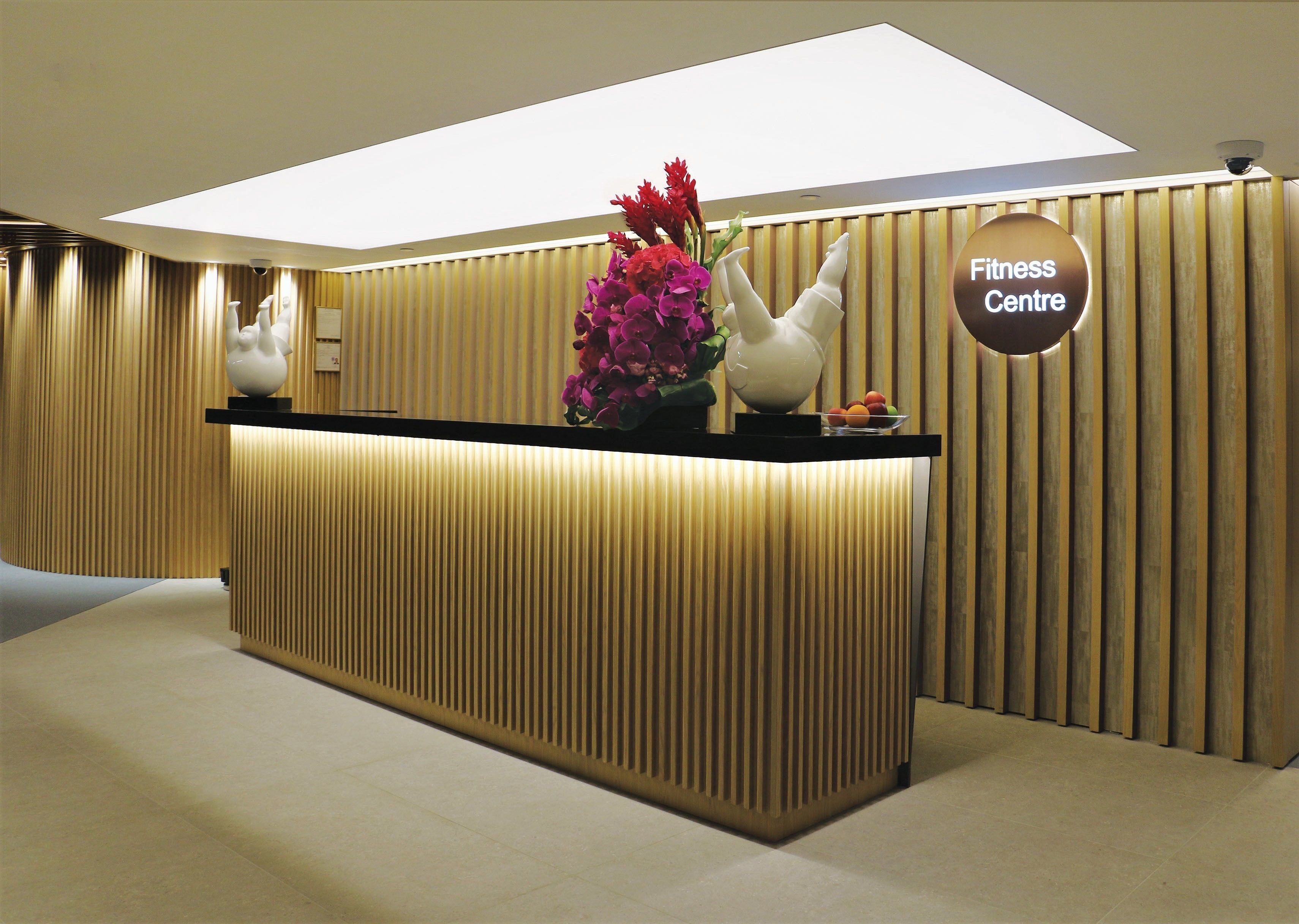 Un mostrador de recepción con un letrero que dice "Fitness Centre" en un interior moderno con paneles de madera verticales y una luz empotrada en el techo.