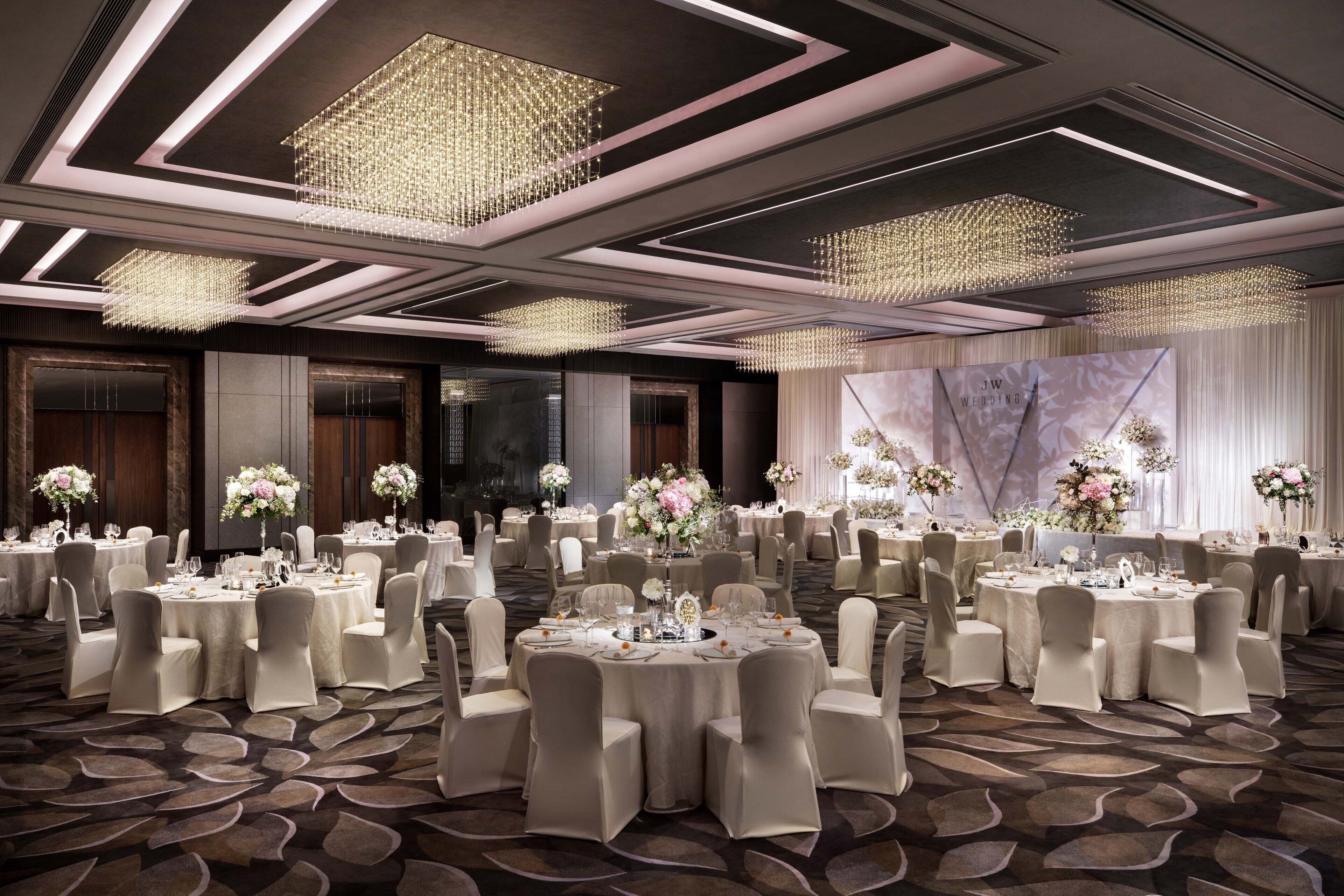 A banquet hall with round tables set for a wedding, featuring chandeliers and floral centerpieces.